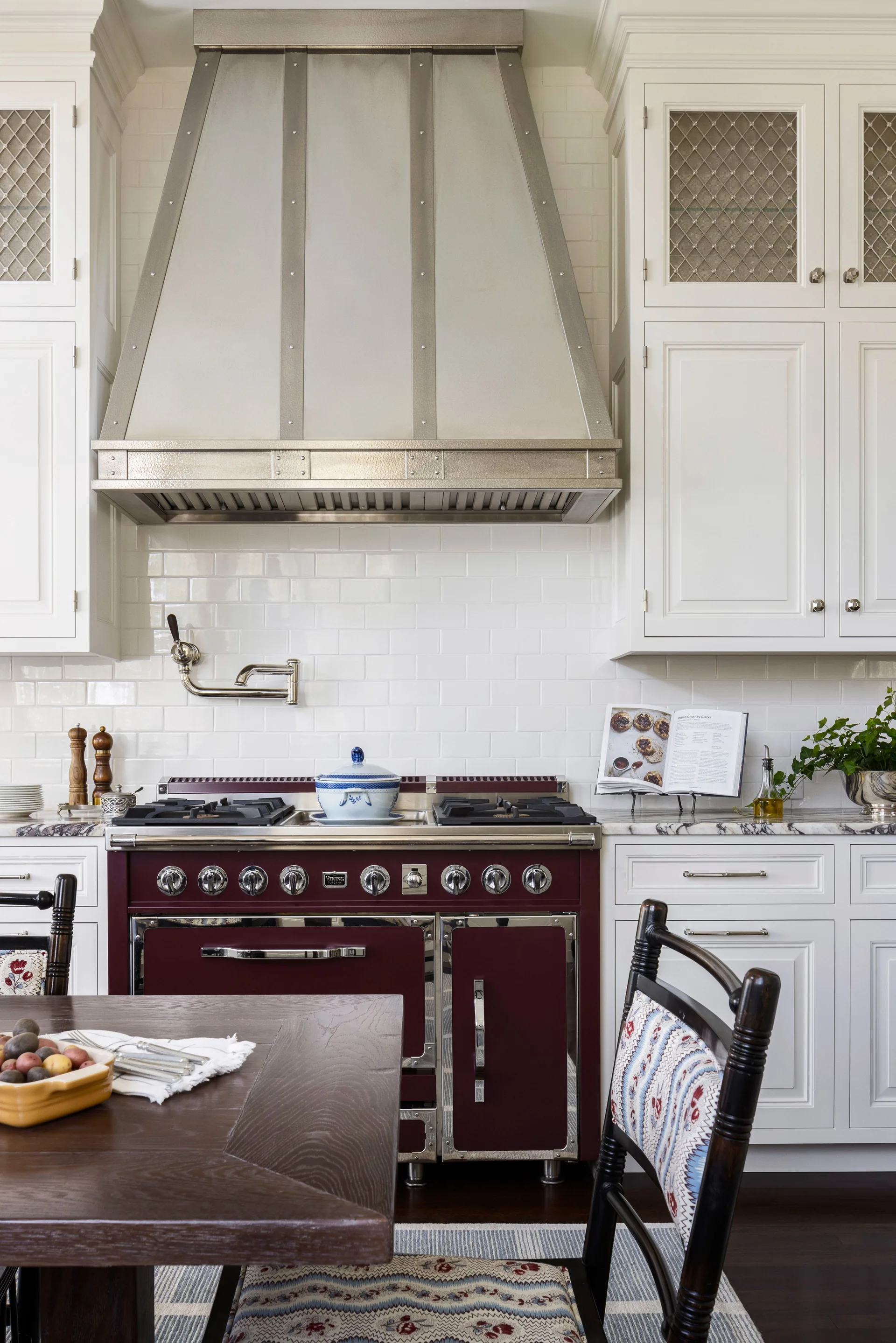 A burgundy kitchen stove sits under a metallic range hood, flanked by white cabinets and accented by a patterned dining chair in the foreground.