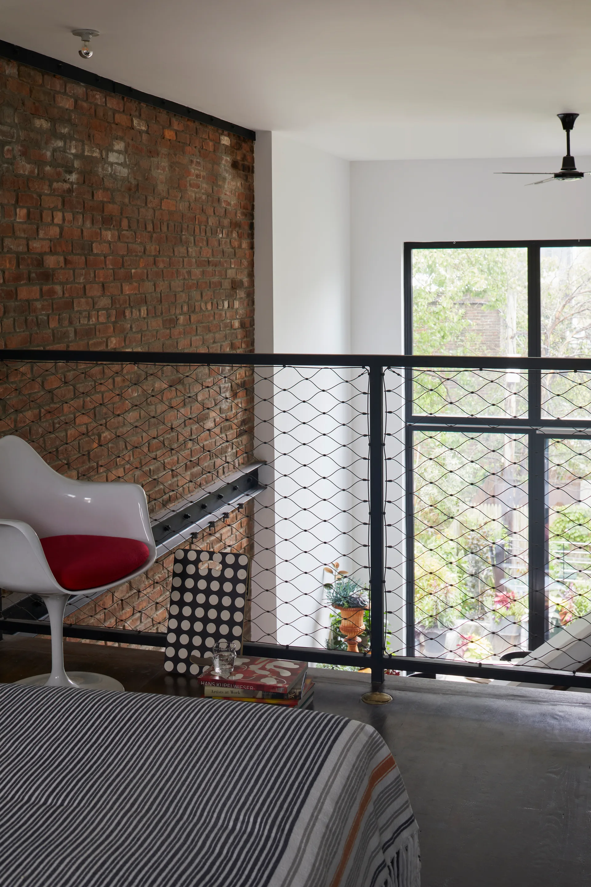 A lofted bedroom features an exposed brick wall, a black metal wire mesh railing, and a white molded chair with a red seat cushion.