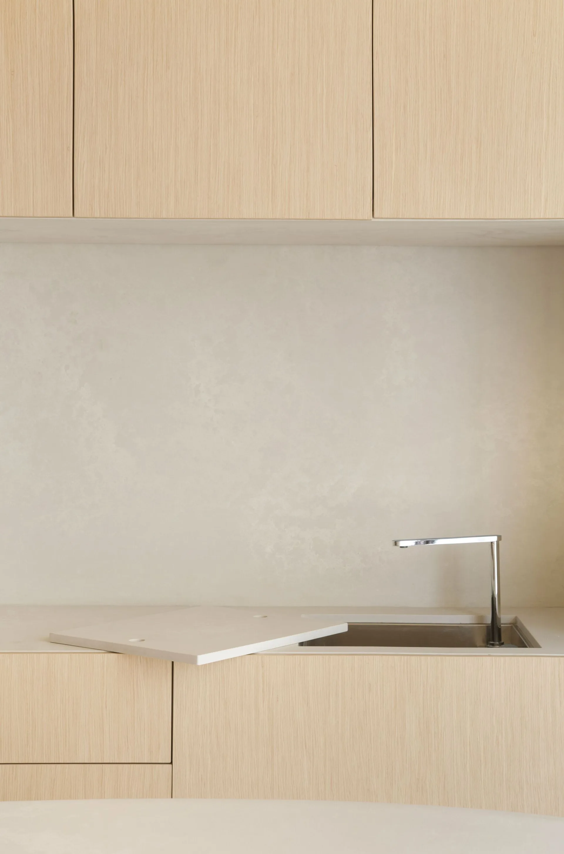 A close-up of a minimalist kitchen counter with light wood cabinets, a textured beige backsplash, and a modern chrome faucet over an integrated sink.