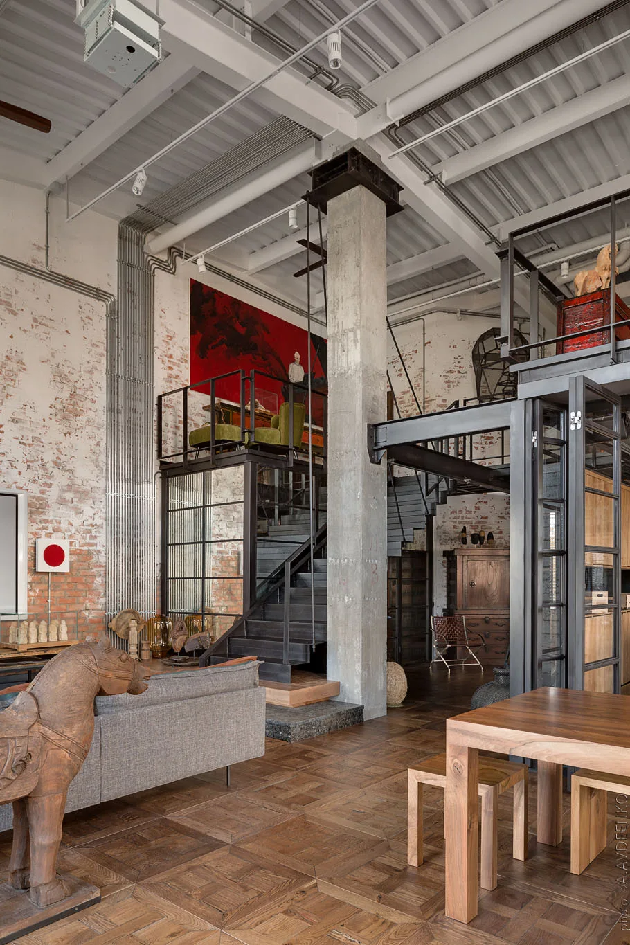 A spacious industrial loft featuring whitewashed brick walls, a central concrete column, and a black steel staircase leading to a mezzanine.