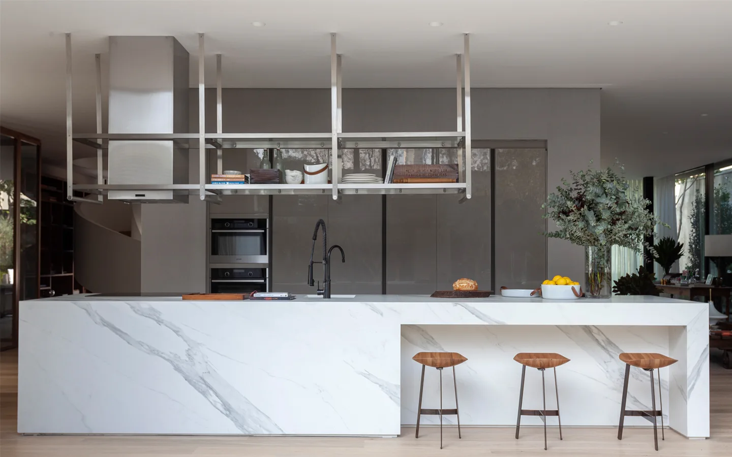 A modern kitchen featuring a white marbled waterfall island with wood bar stools and an overhead stainless steel shelving unit against gray cabinets.