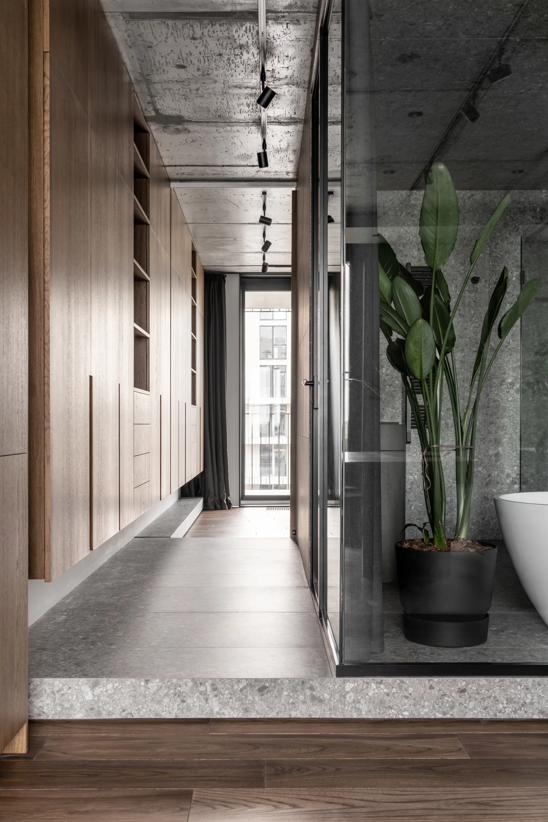 A modern hallway features floor-to-ceiling wood cabinetry, a raw concrete ceiling, and a glass-enclosed bathroom containing a freestanding tub.