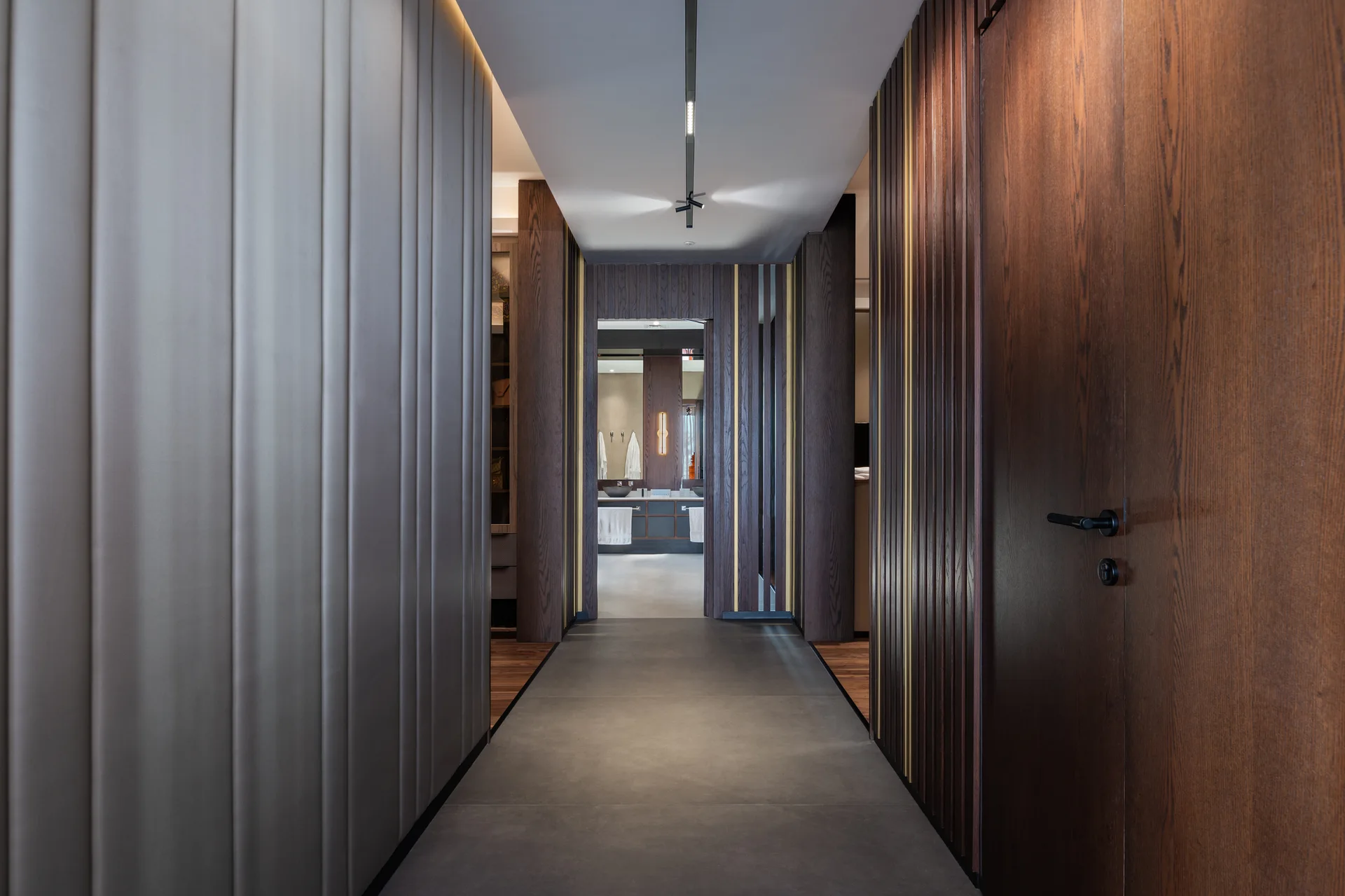 A modern hallway features dark vertical wood slats and grey wall panels leading to a brightly lit bathroom doorway.