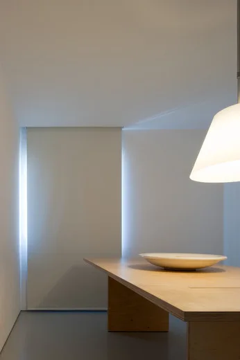A minimalist dining room features a long wooden table with a white ceramic bowl under a pendant light and recessed vertical wall lighting.