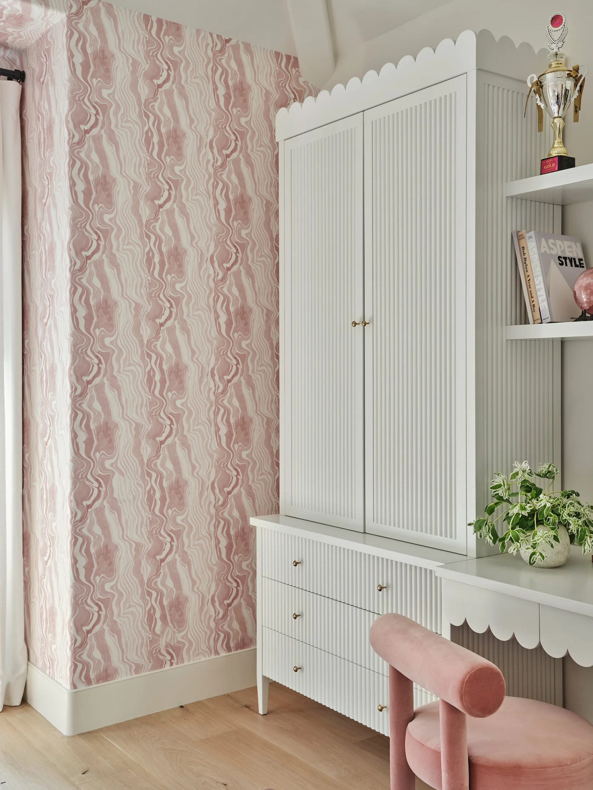 A tall white fluted cabinet with scalloped trim stands against a pink marbled wallpaper wall next to a matching desk and a pink chair.