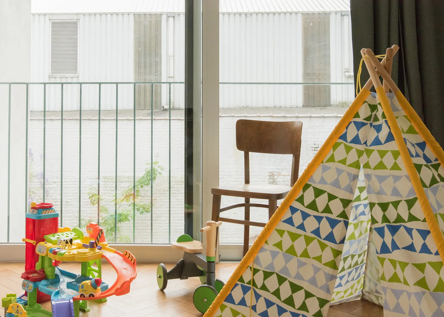 A fabric play teepee with a green and blue geometric pattern stands on a herringbone parquet floor next to a wooden chair and plastic toys.