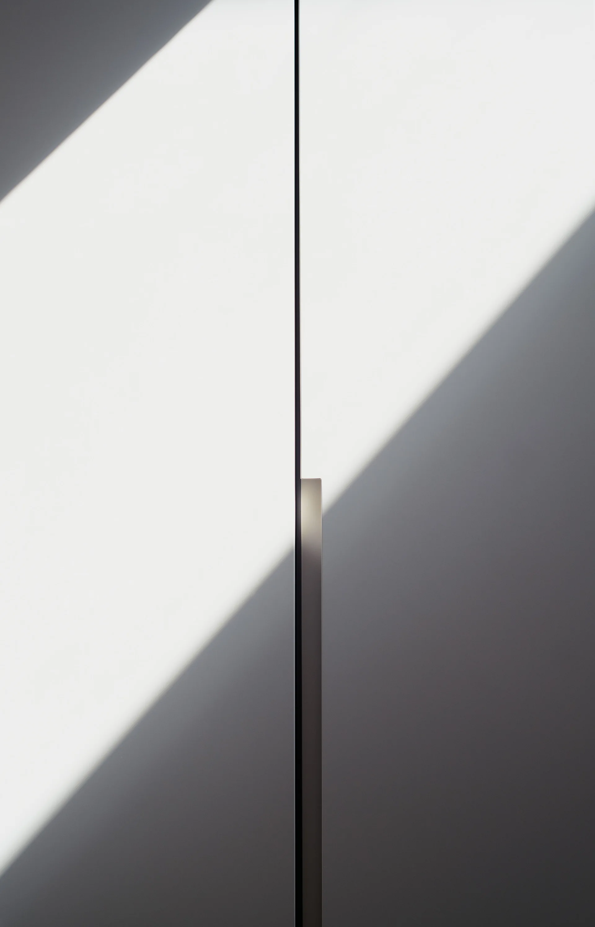 A close-up of matte white cabinet doors with a slim vertical metal handle and a sharp diagonal shadow casting across the surface.