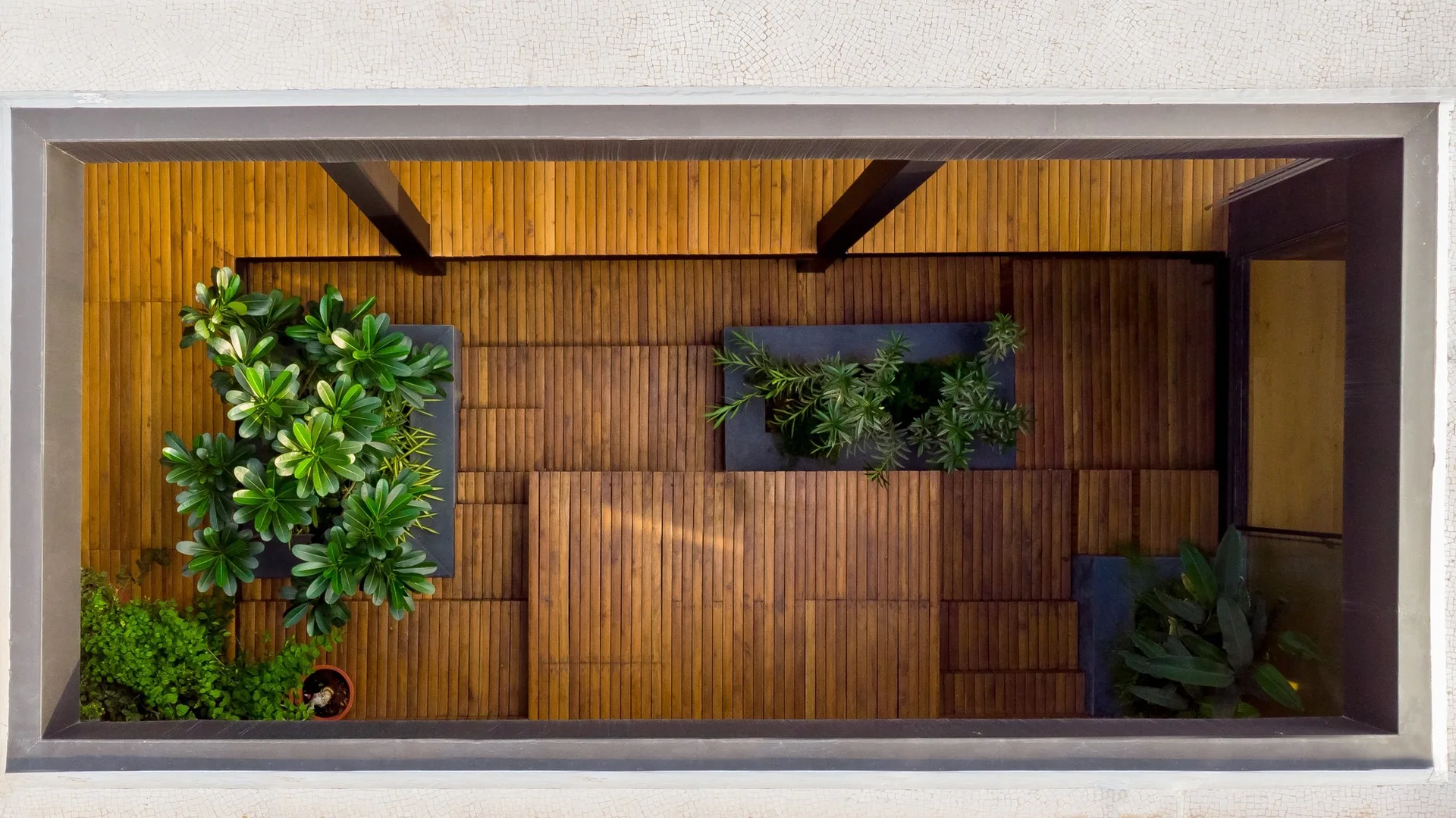 An overhead view of a modern courtyard with brown slatted wood decking and several rectangular built-in dark gray planters holding green plants.
