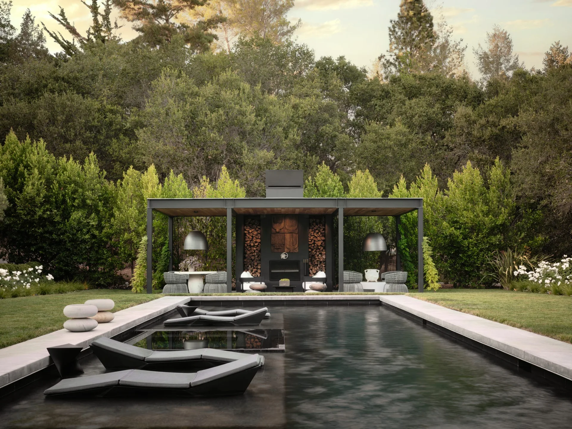 A dark-bottomed swimming pool with integrated lounge chairs sits before a modern black metal pavilion featuring a stone fireplace and woven outdoor seating.