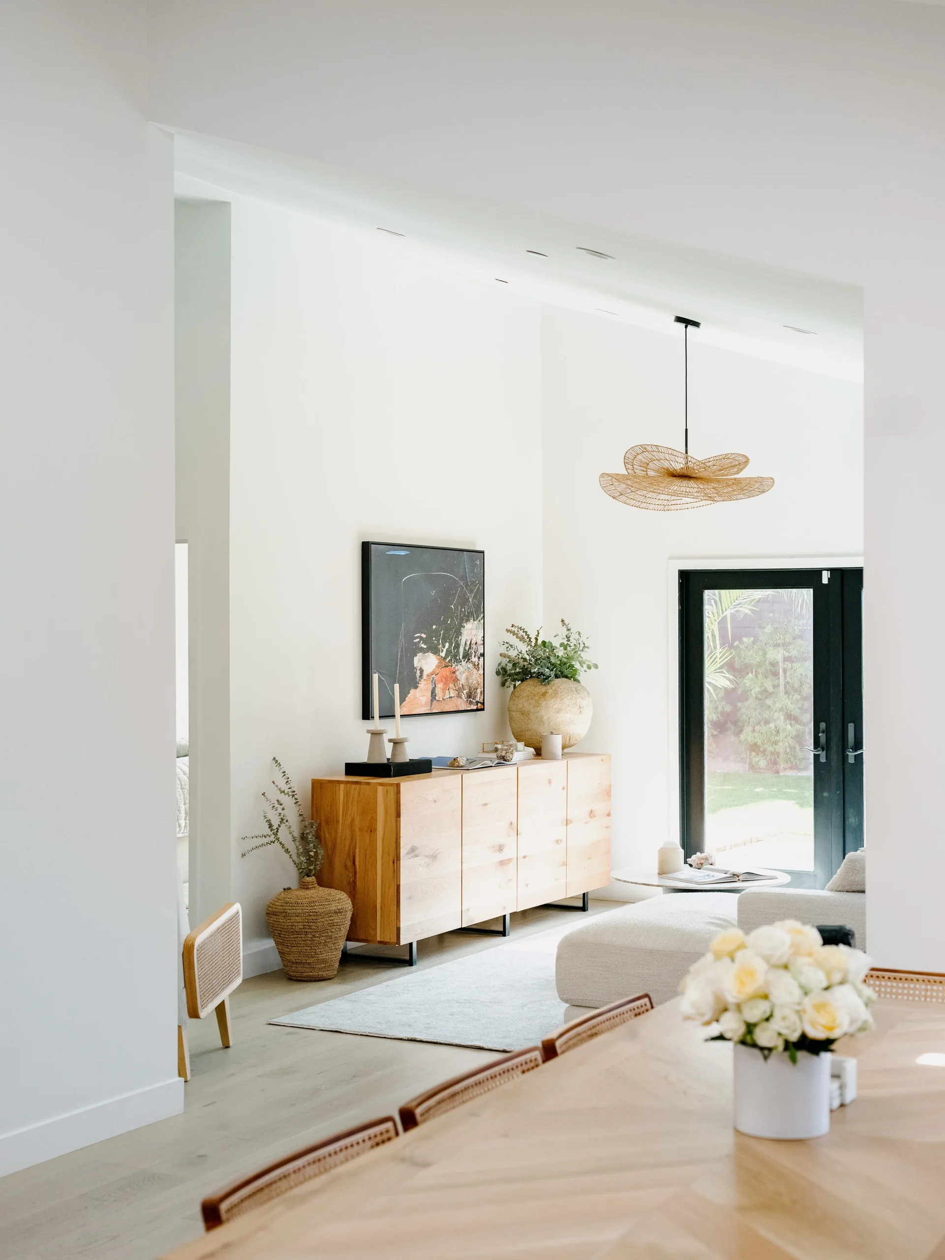 A bright living room features a natural wood sideboard with decor, a white sofa, and a woven pendant light hanging from a vaulted ceiling.