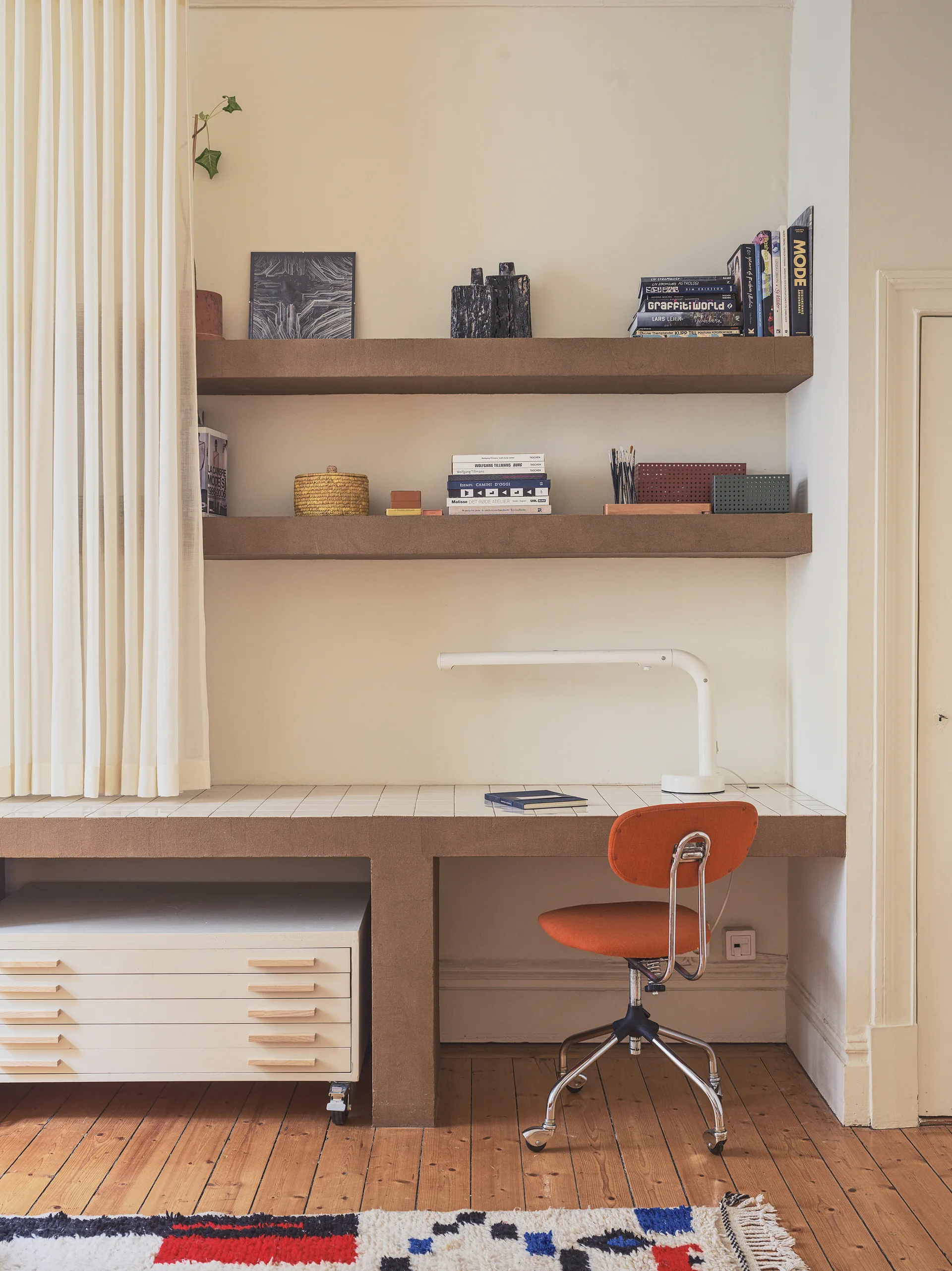 A home office workspace features a brown built-in desk with a white tiled top, an orange rolling task chair, and matching brown floating shelves.