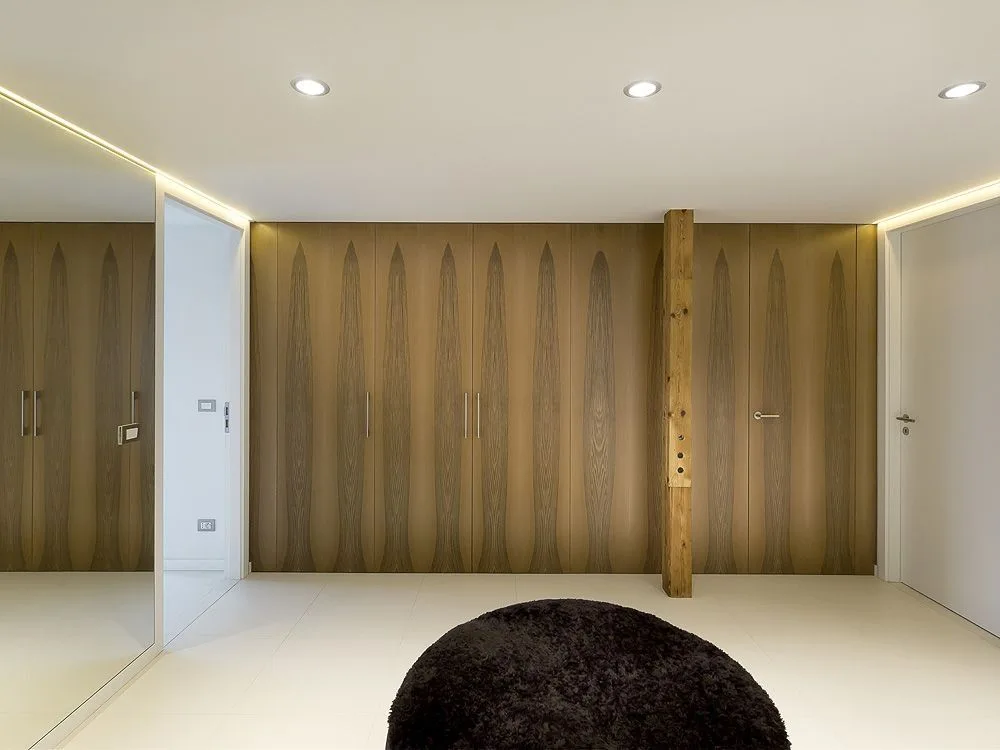 A minimalist hallway features a wall of wood-grained cabinets, a rustic wooden column, a mirrored wall, and a black circular rug.