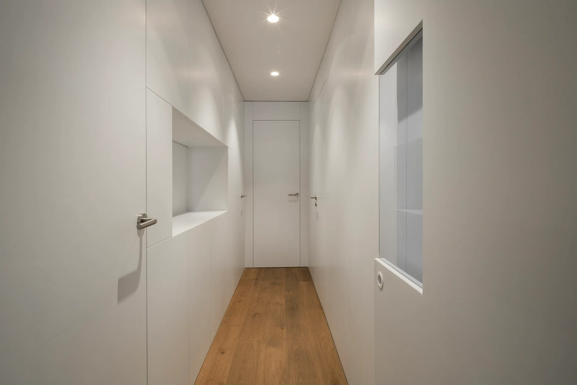 A long minimalist hallway features seamless white wall panels with flush doors, a recessed wall niche, and natural wood plank flooring.