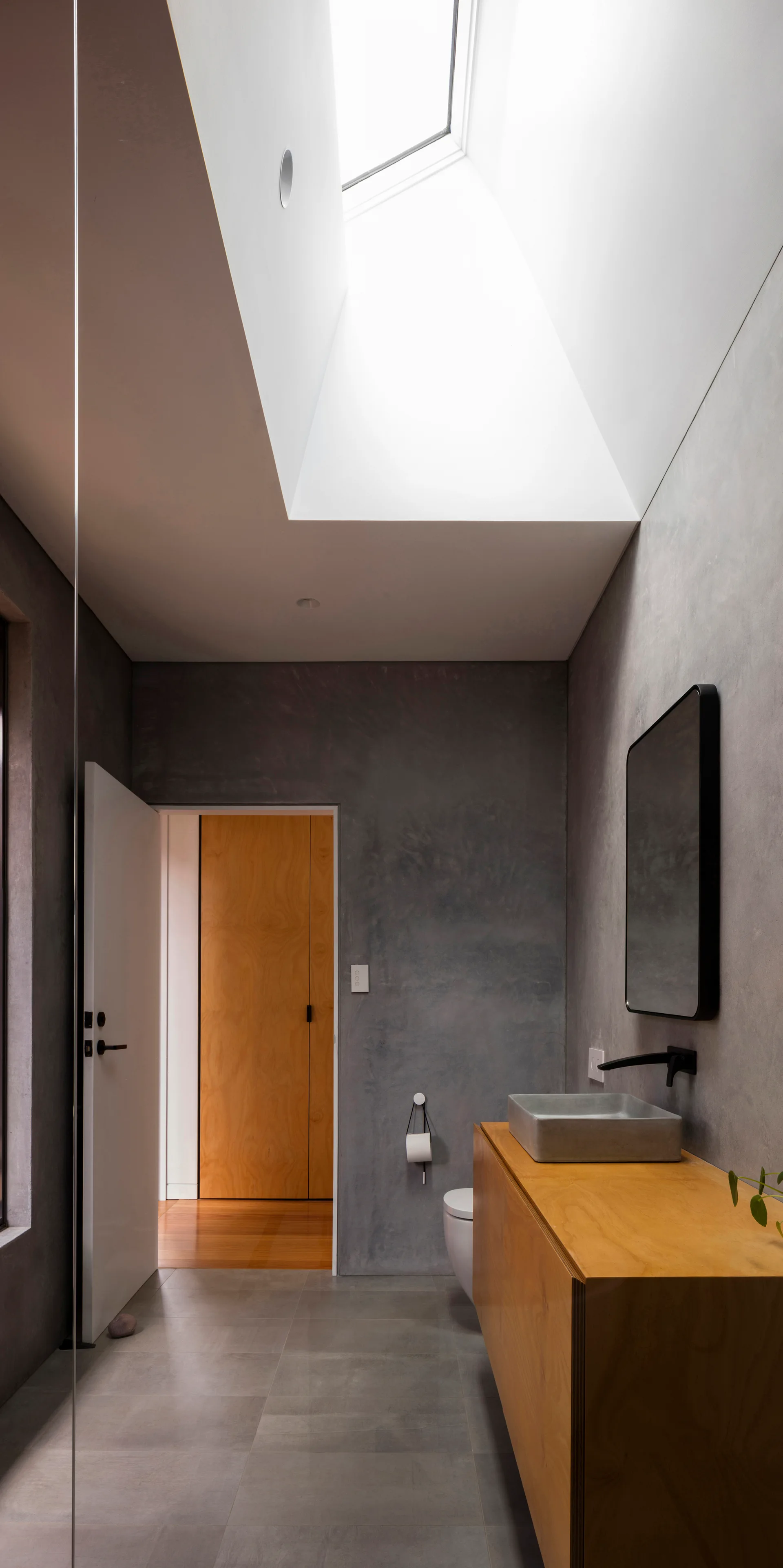 A minimalist grey bathroom featuring a light wood vanity, a rectangular concrete vessel sink, and a large angled skylight shaft.
