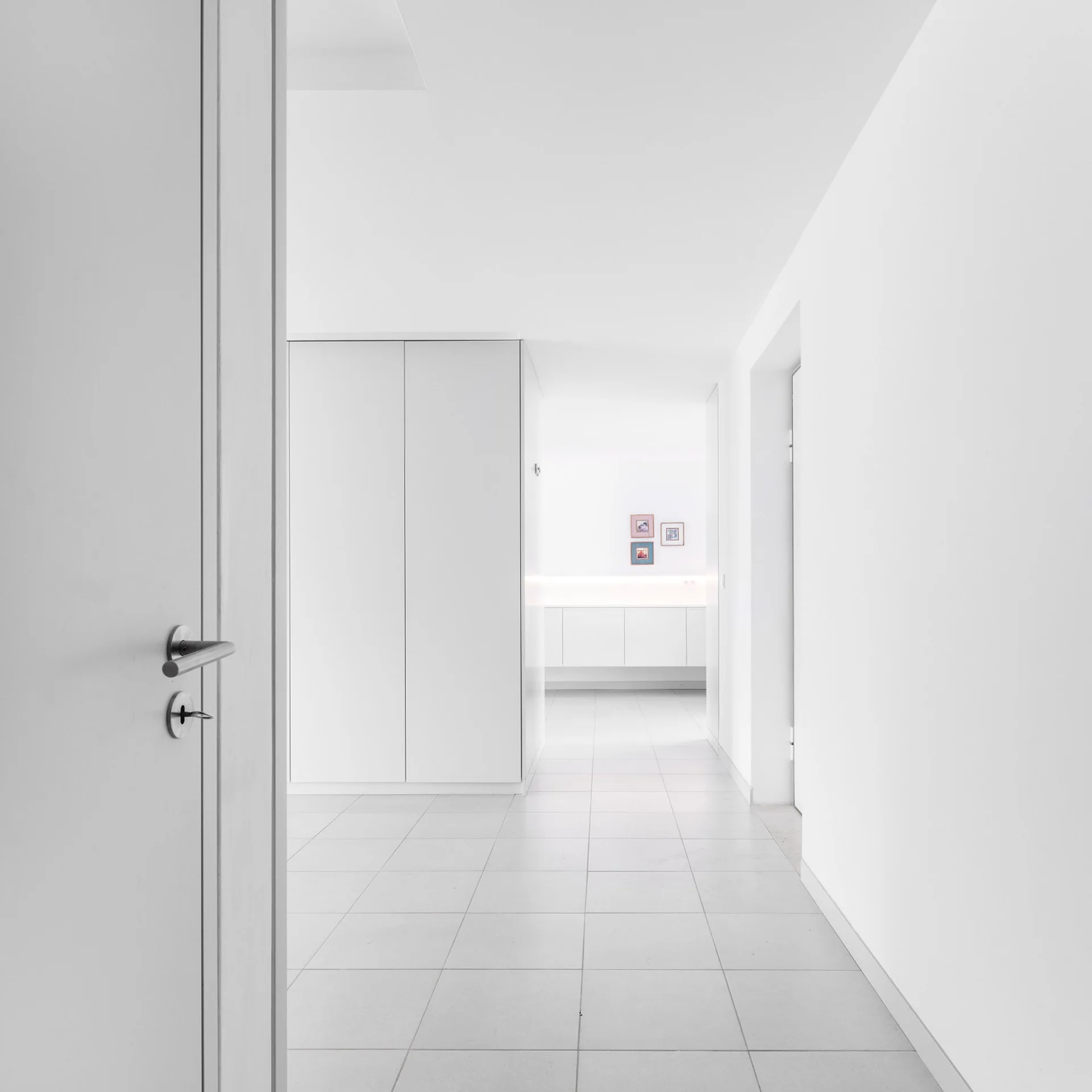 A view through an open door into a minimalist white hallway featuring large square floor tiles, flush built-in cabinets, and warm under-cabinet lighting.