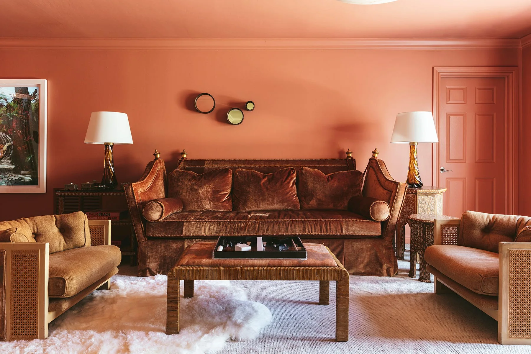 A living room with terracotta walls and ceiling features a brown velvet sofa, two cane armchairs, and matching amber glass table lamps.