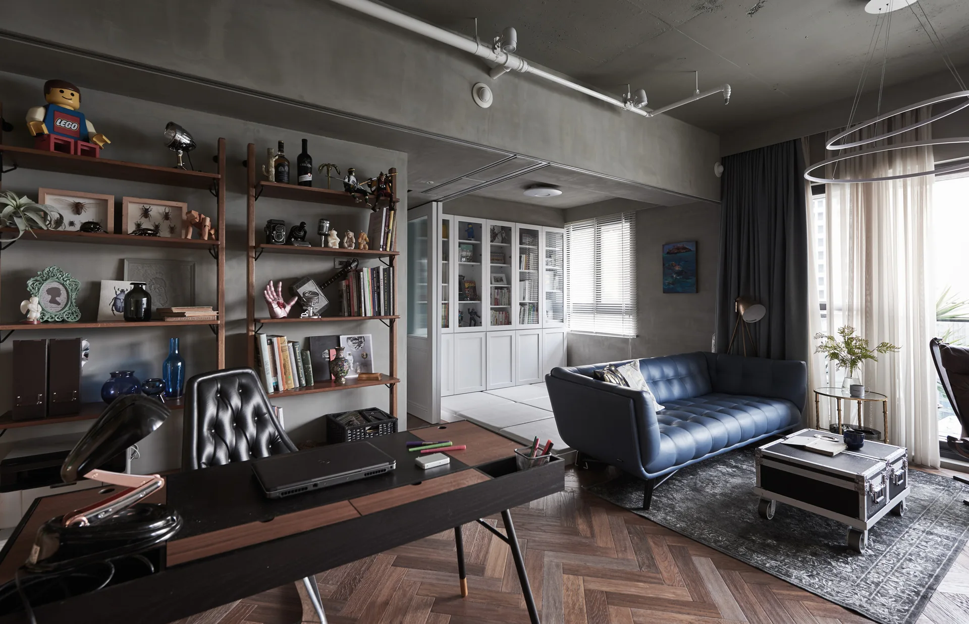 A combined industrial living room and workspace features a blue leather sofa, a dark wood desk, concrete walls, and herringbone pattern wood flooring.