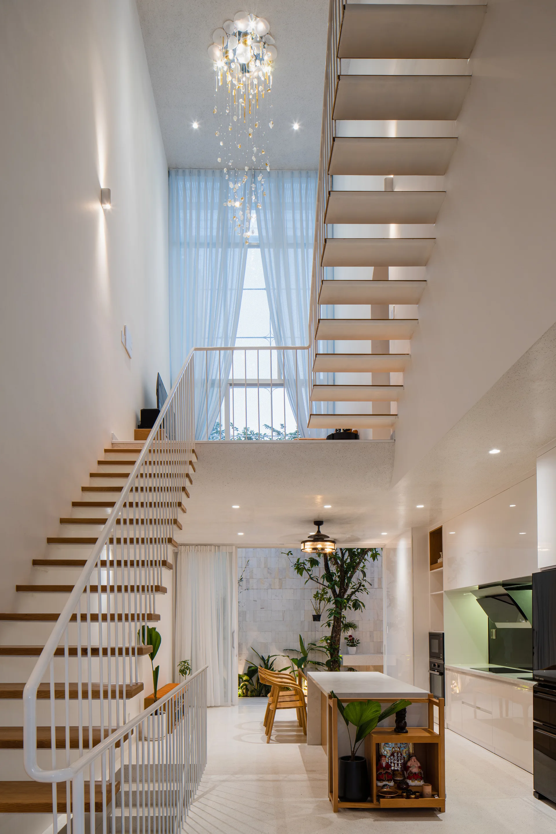A double-height modern interior featuring a floating white staircase, a long glass chandelier, and a white kitchen with a rear courtyard view.