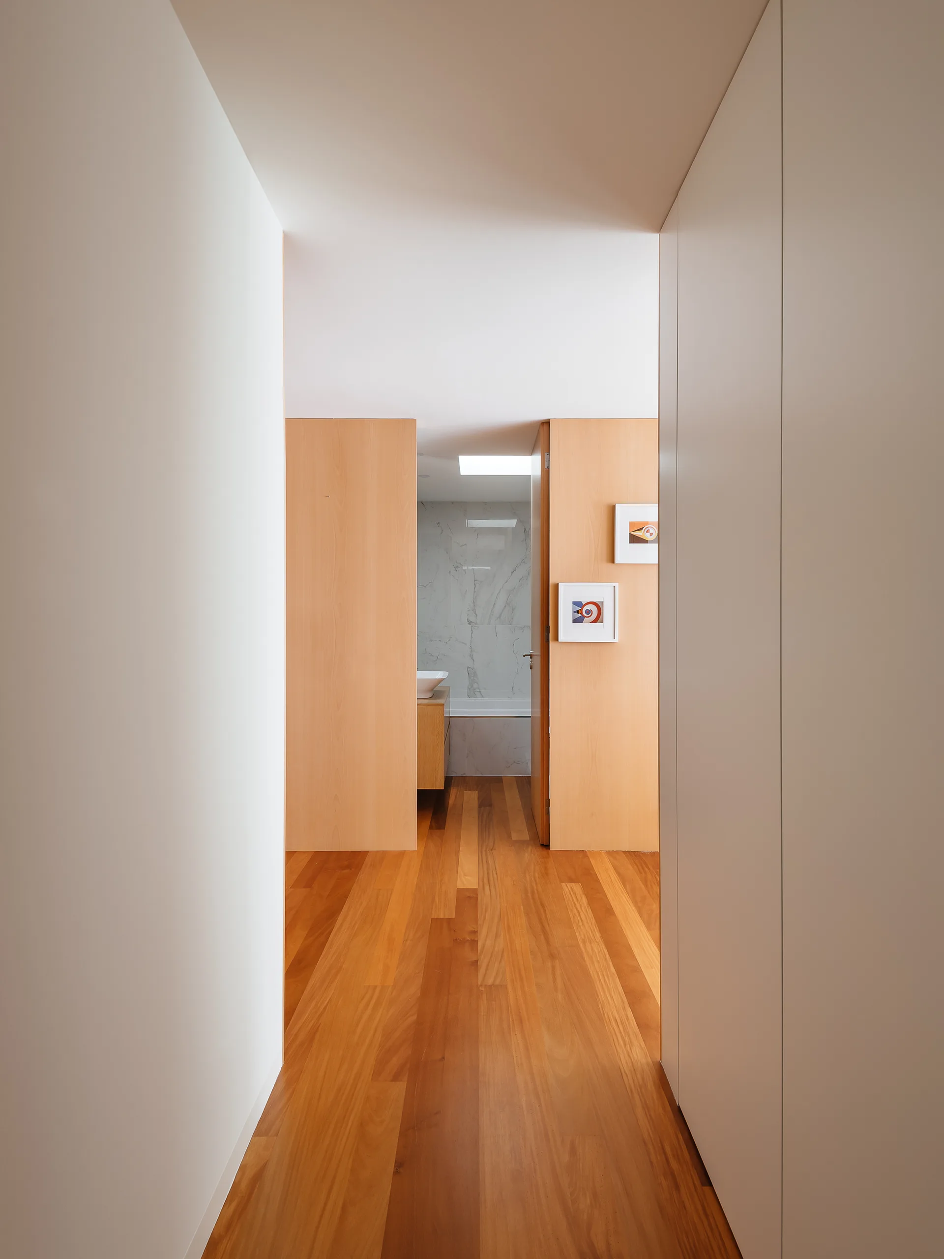 A minimalist hallway with warm hardwood floors leading past a light wood partition wall toward a bathroom with marble tiling.