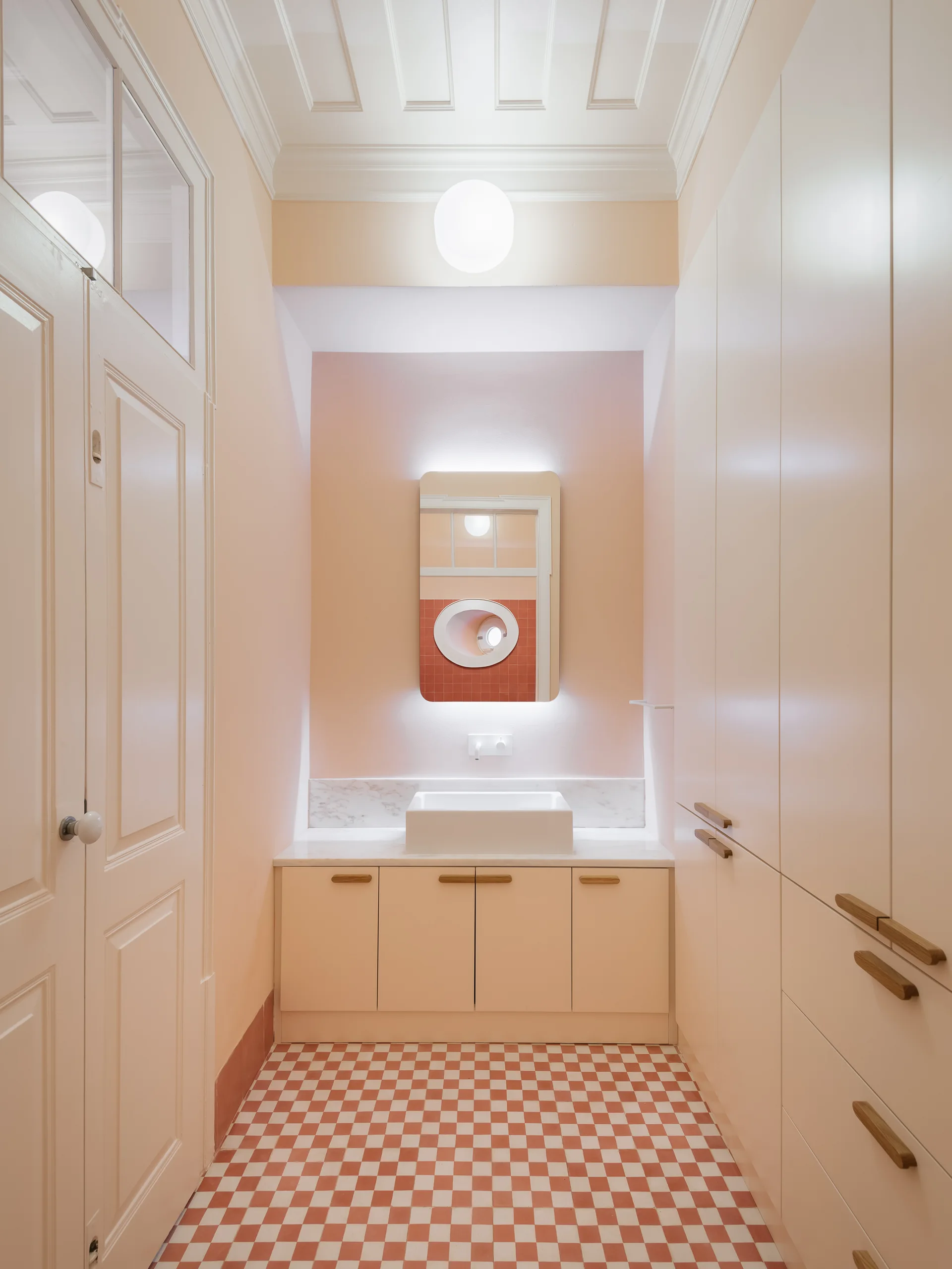 A narrow pink bathroom features a backlit rectangular mirror over a stone vanity, custom off-white cabinetry, and a terracotta checkered tile floor.
