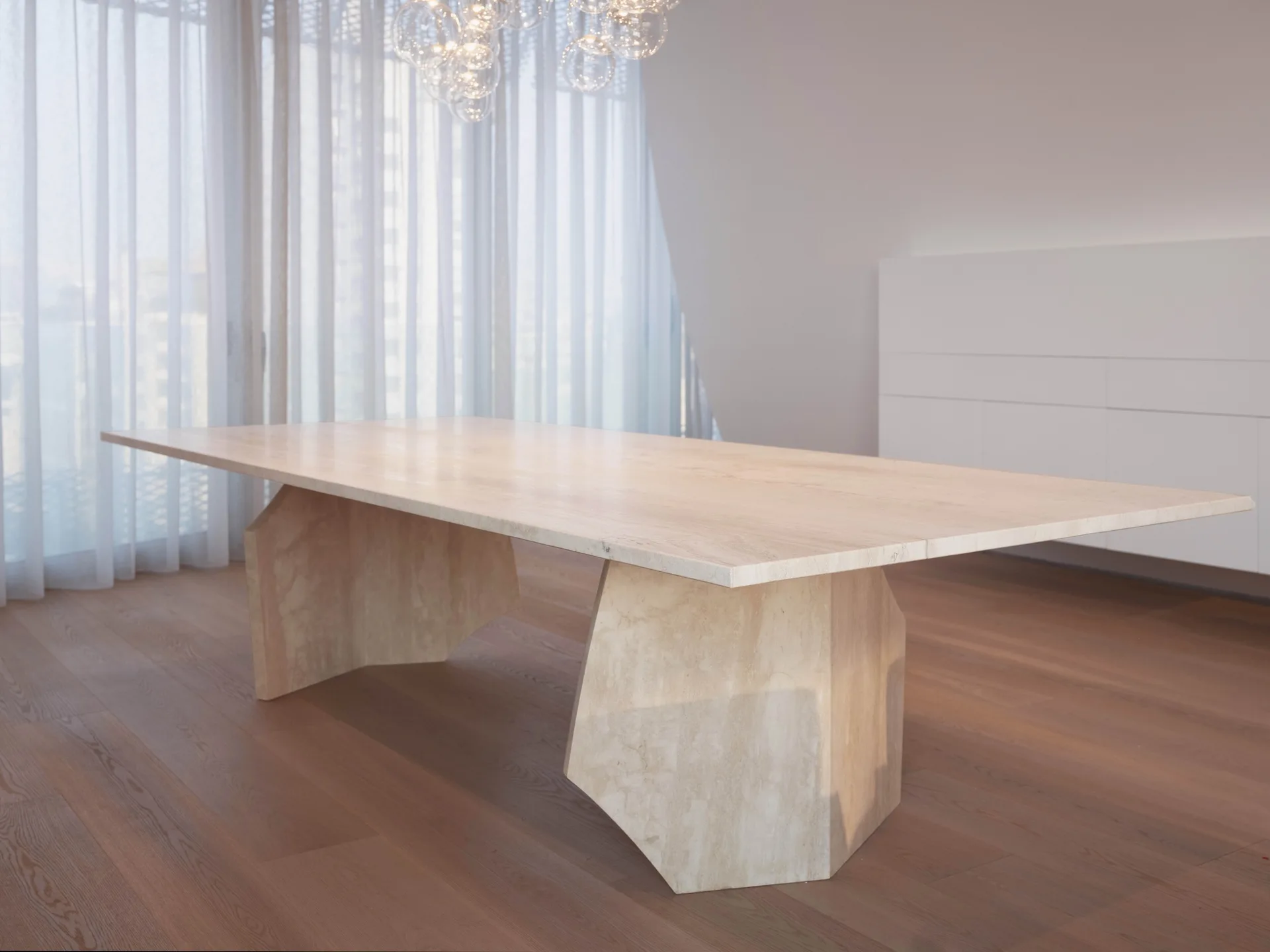 A large geometric stone dining table sits on wood flooring in a minimalist dining room with white walls and floor-to-ceiling sheer curtains.