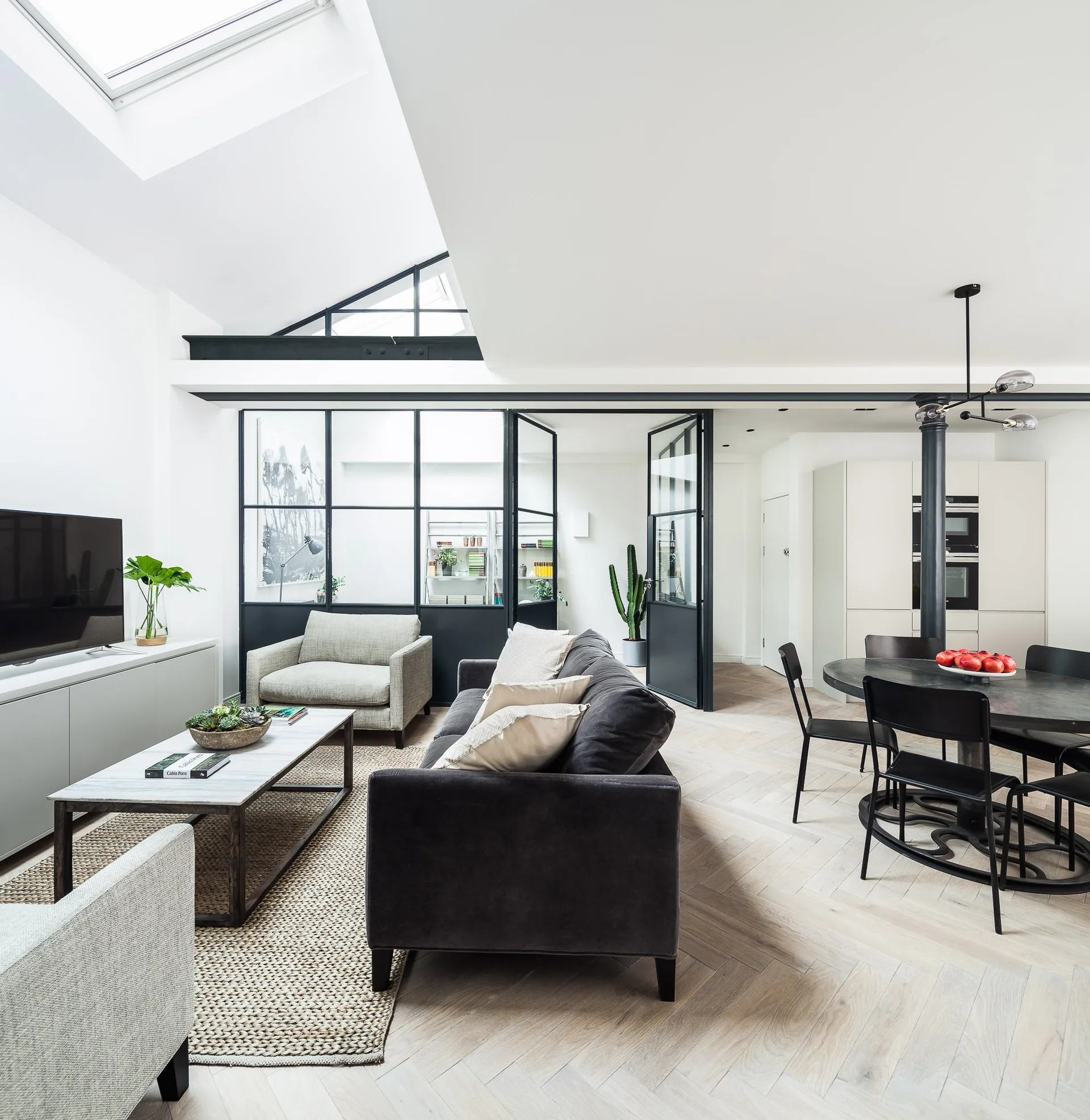 A modern open-concept living and dining area features a dark sofa, light wood herringbone flooring, and a black-framed glass partition wall.