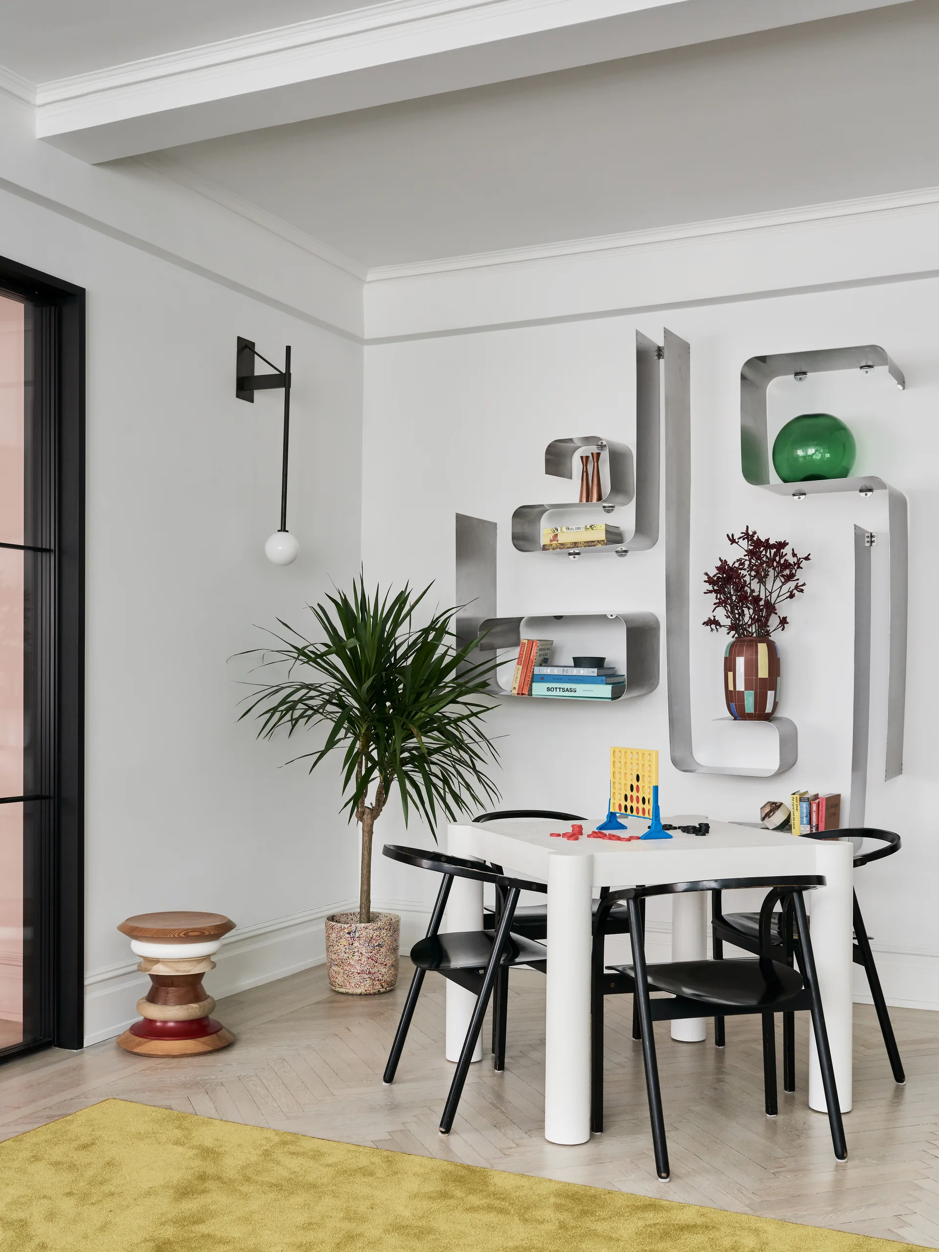 A white square dining table with black chairs sits in a room featuring winding metal wall shelves, a tall potted plant, and herringbone wood floors.