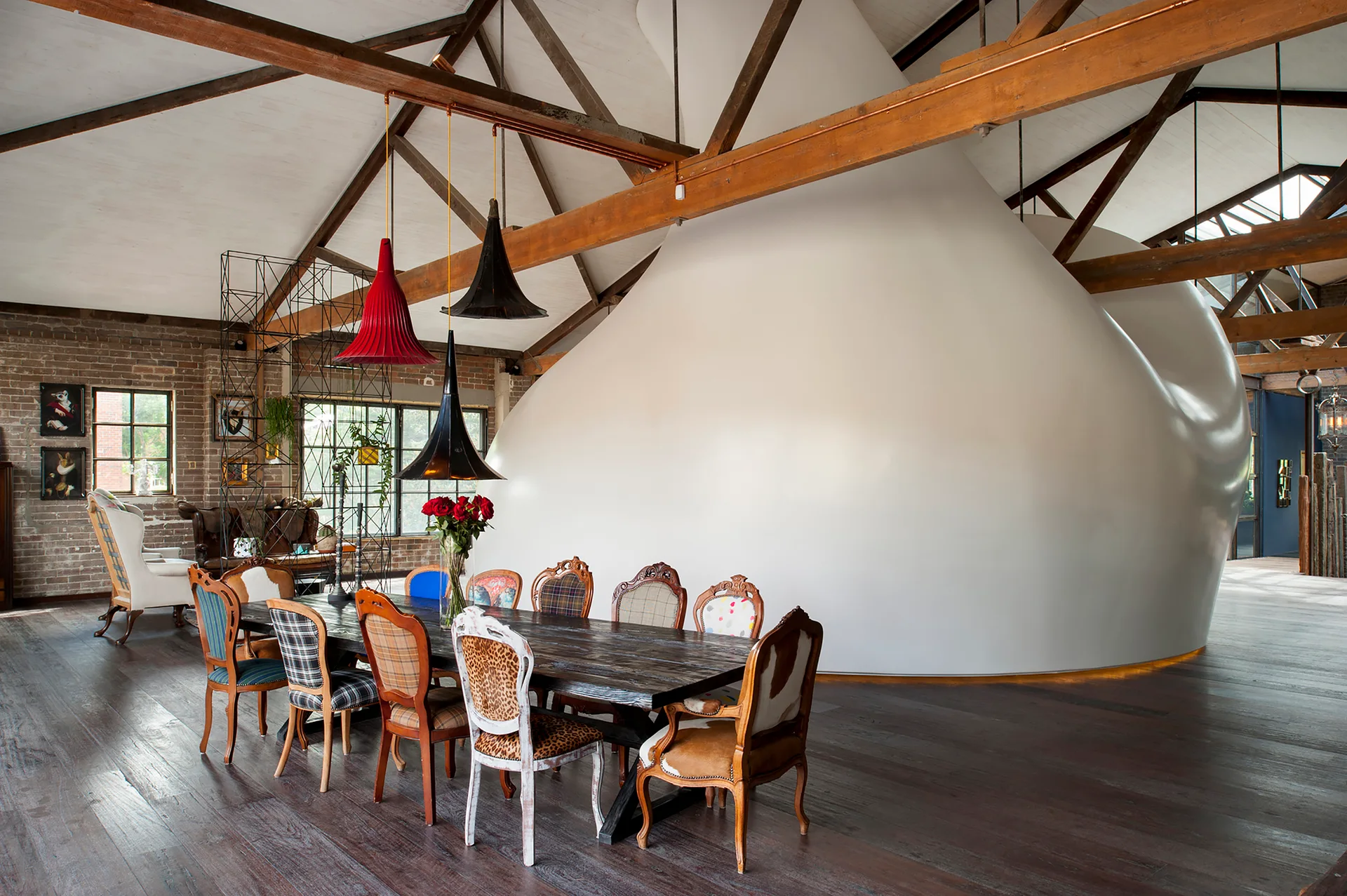 A dark wood dining table surrounded by mismatched patterned chairs sits beneath exposed timber roof trusses in an open industrial loft.
