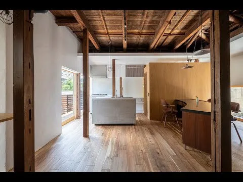 An open-plan kitchen and workspace features a concrete island, exposed timber ceiling beams, wide-plank wood flooring, and a curved desk.
