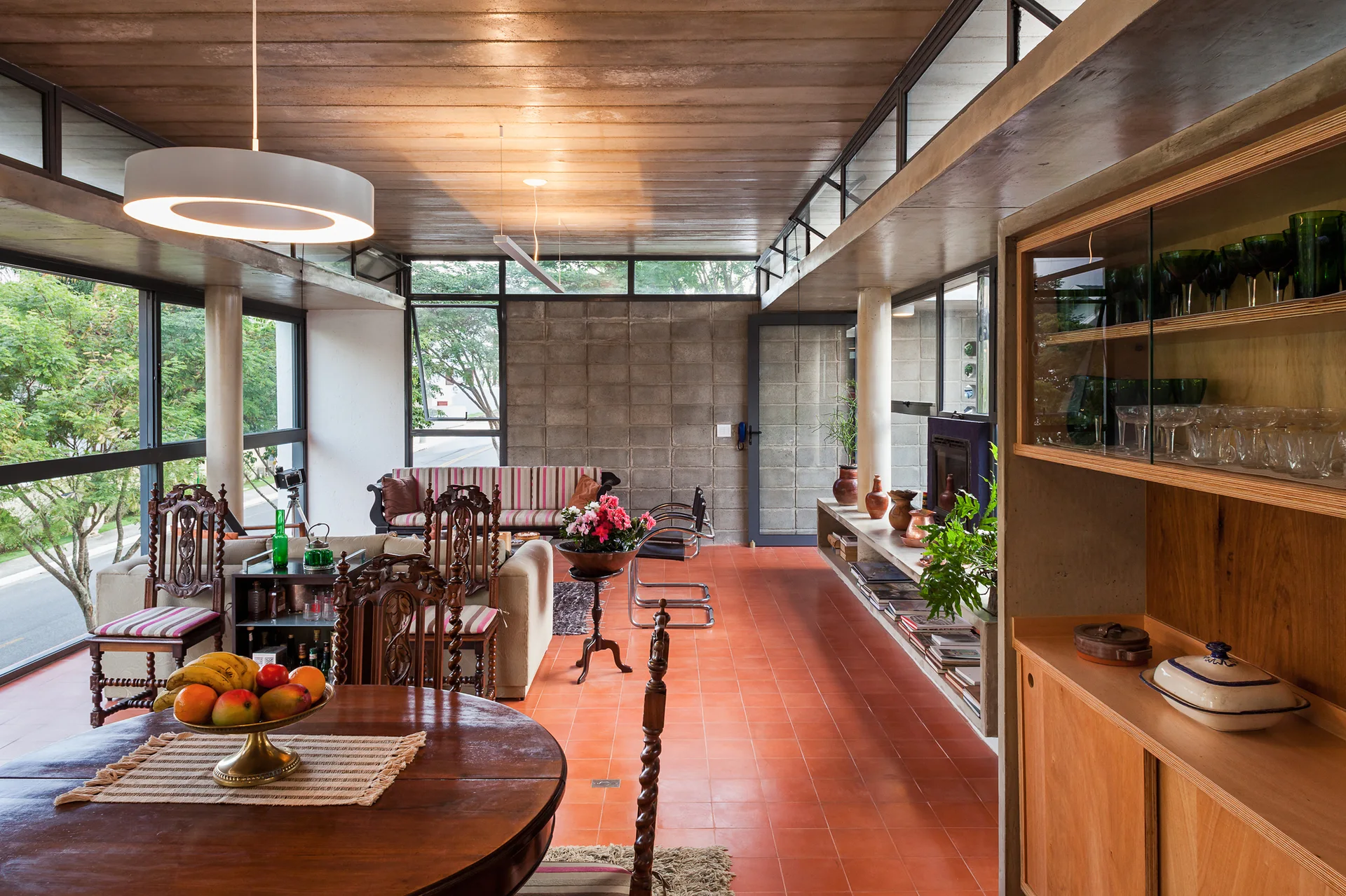 An open-plan living and dining area features terracotta floor tiles, a grey concrete block wall, a wood ceiling, and mid-century furniture.