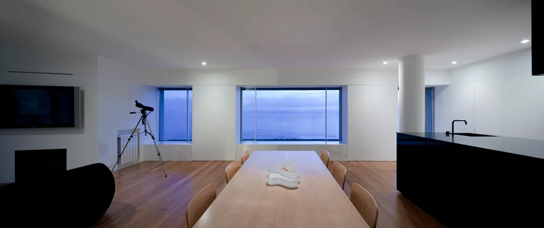 A minimalist dining room features a long light wood table, a black kitchen island, and wide windows displaying a blue ocean view.