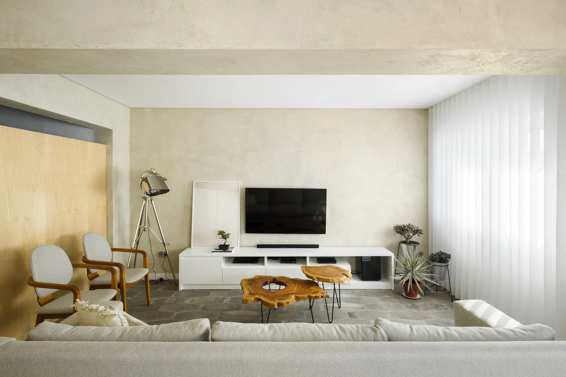 A minimalist living room features a white media console beneath a wall-mounted TV, twin live-edge wood coffee tables, and textured beige plaster walls.