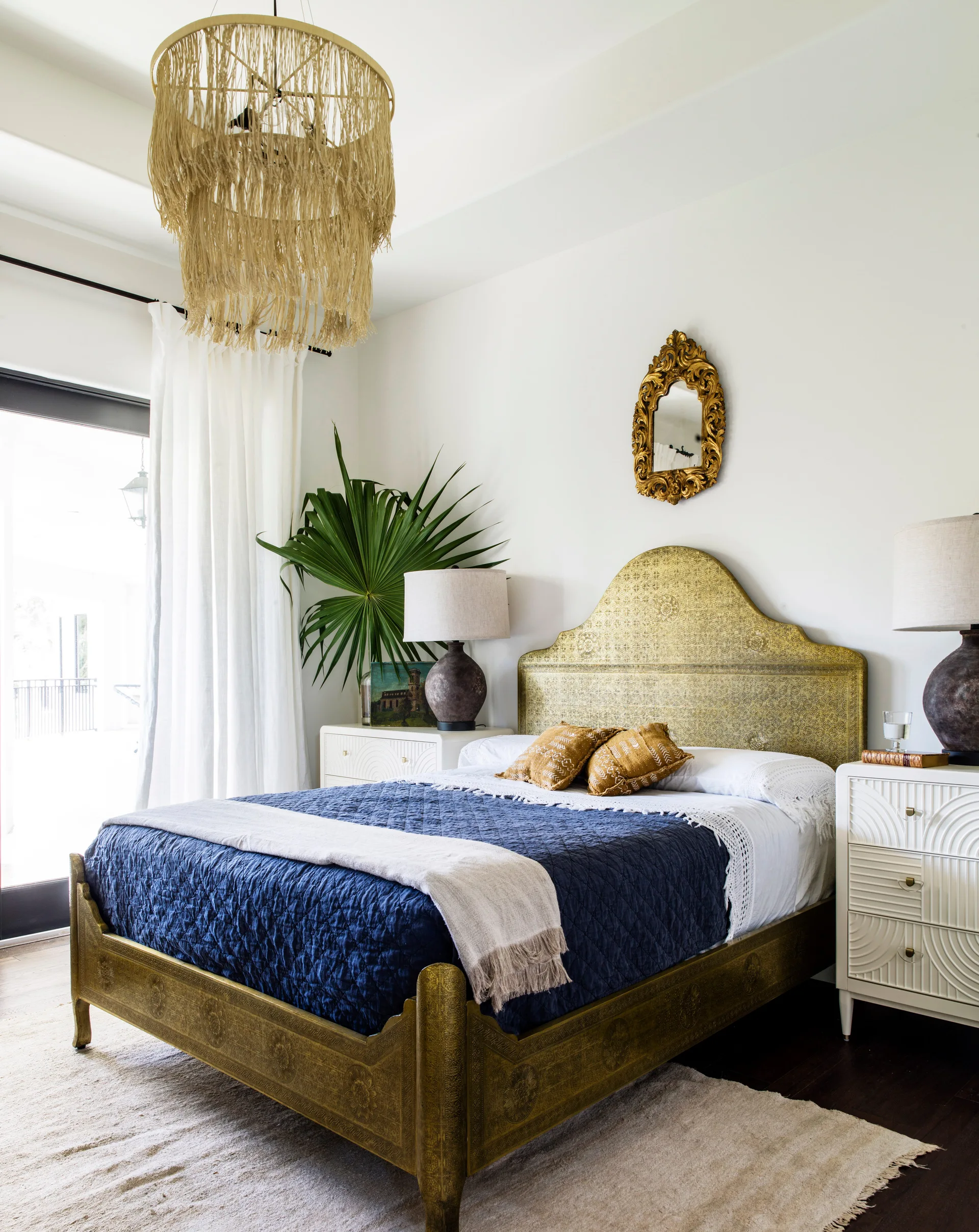 An eclectic bedroom features an engraved gold bed frame, white textured nightstands, a navy blue quilt, and a hanging fringed chandelier.