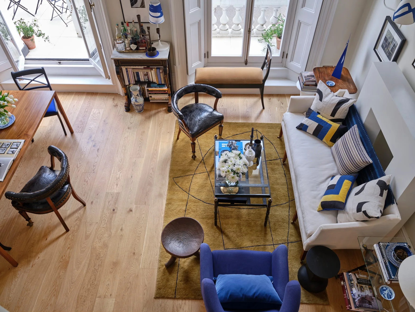 An overhead view of an eclectic living room with a white sofa, a mustard yellow geometric rug, a blue armchair, and light hardwood floors.