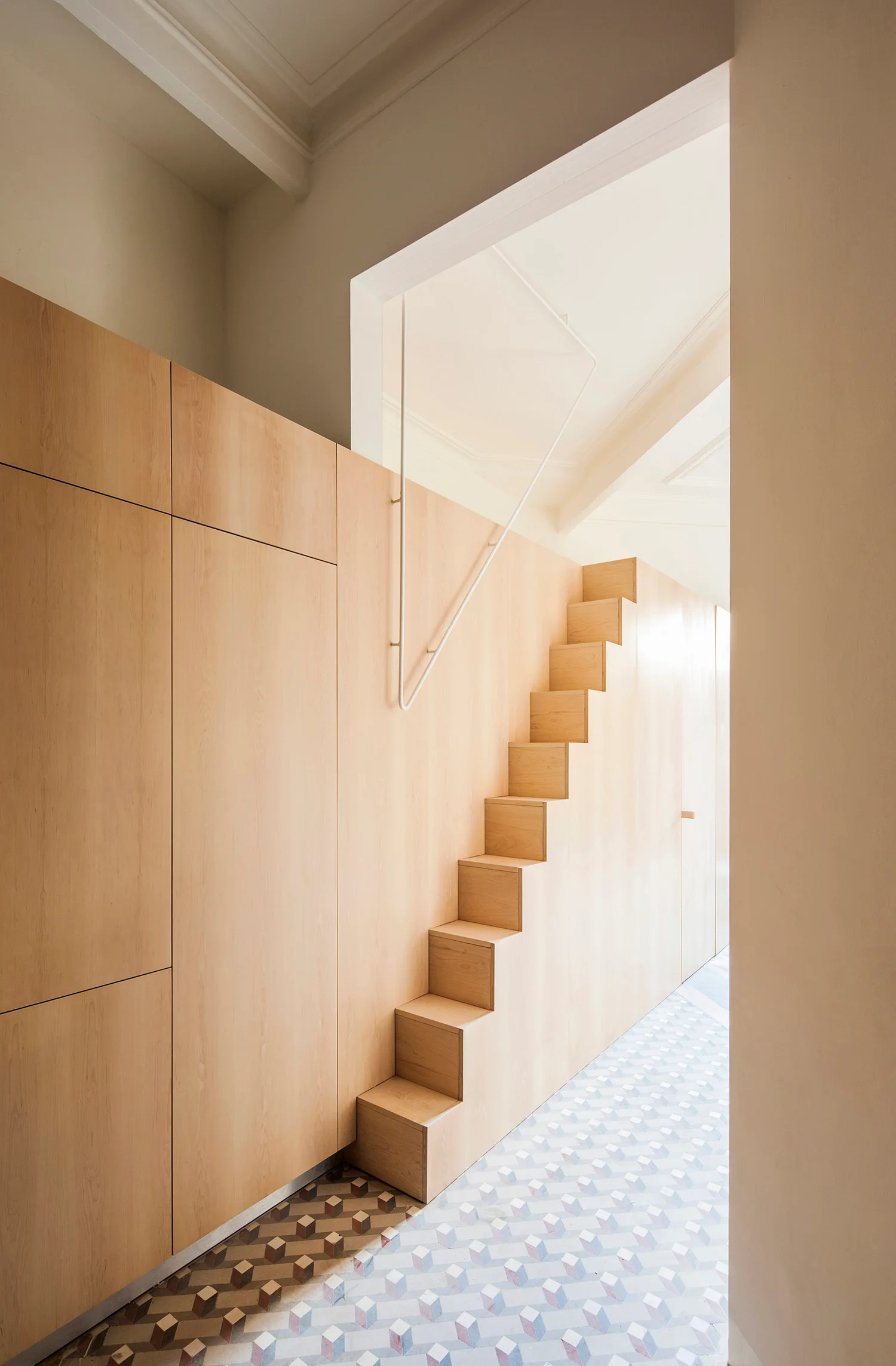 A light wood staircase integrated into a floor to ceiling cabinet wall above a geometric cube patterned tile floor.