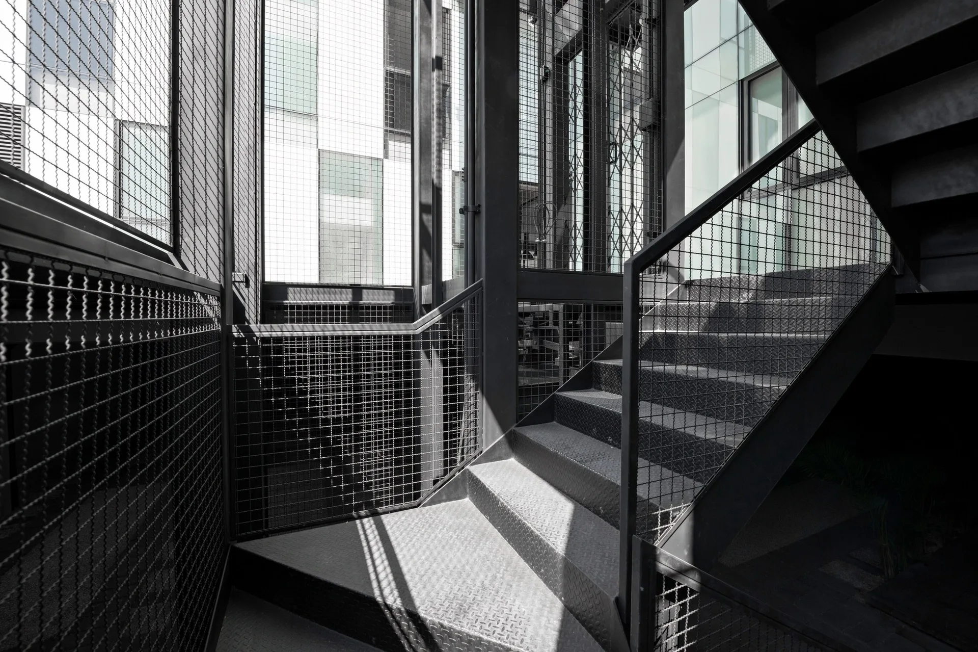 An industrial black metal staircase features diamond plate treads and woven wire mesh safety railings under bright natural sunlight.