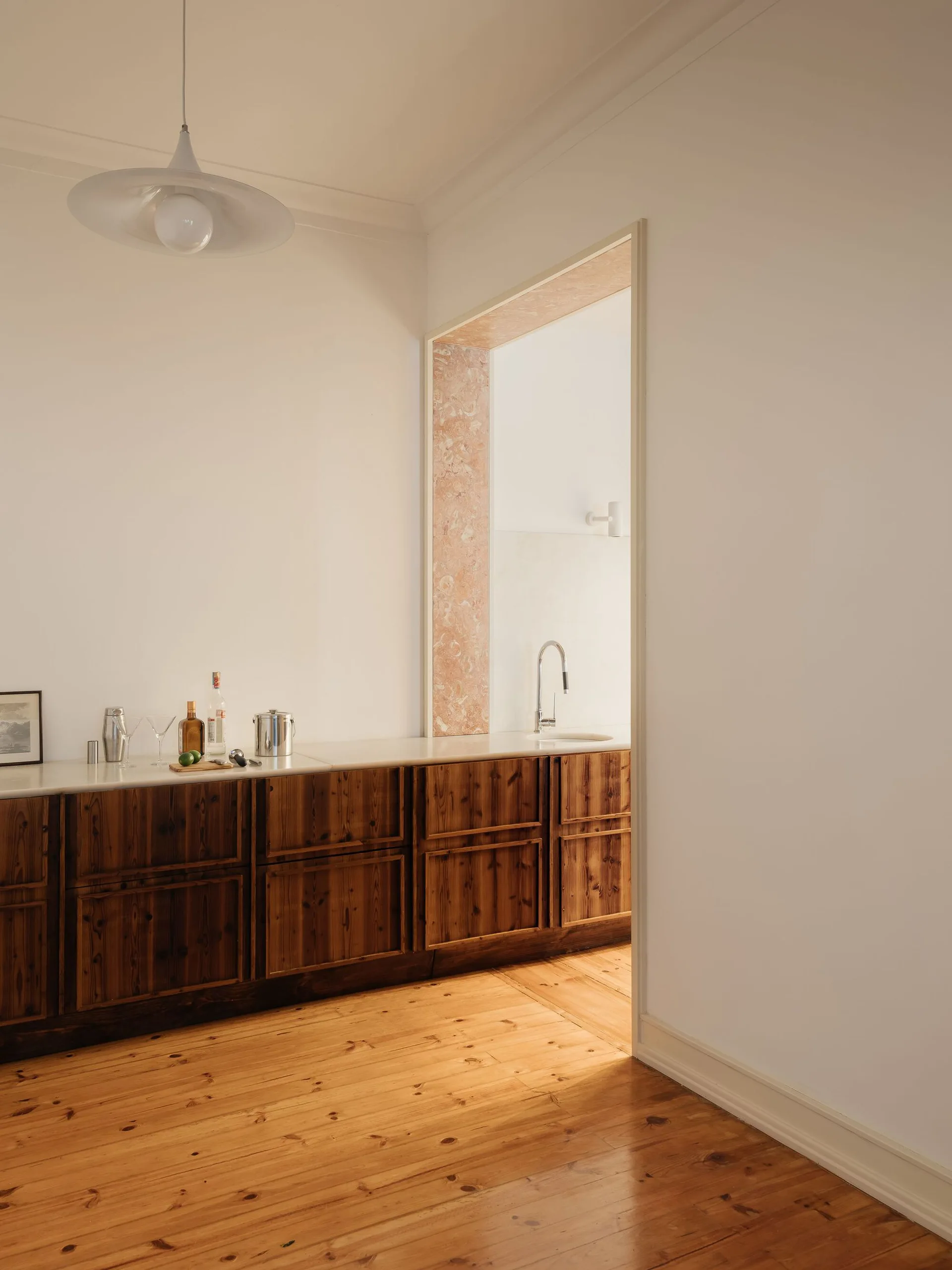 A minimalist kitchen features long dark wood base cabinets under a white countertop, light wood flooring, and a wide doorway framed with pink stone trim.