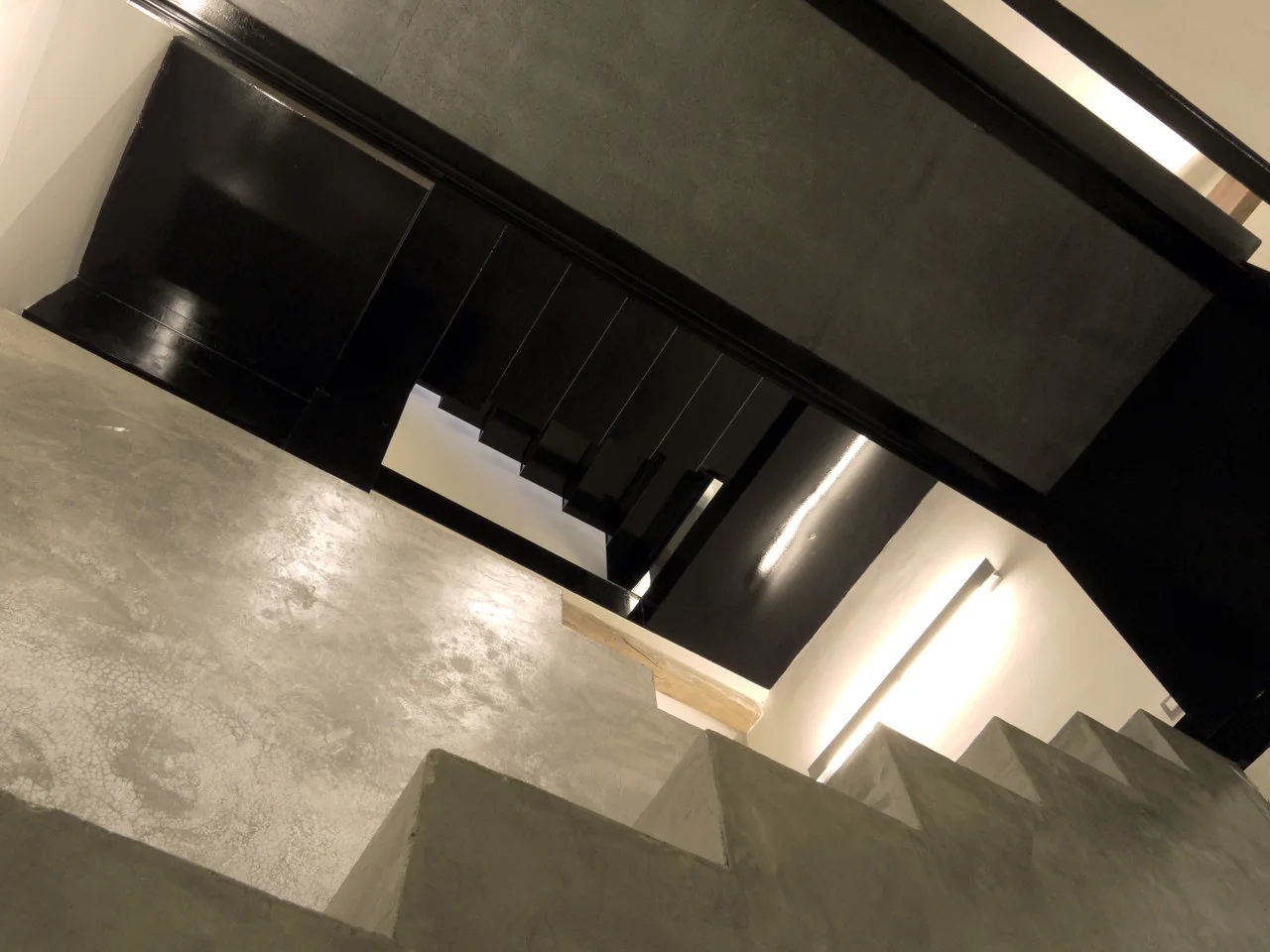 An angled view of a modern industrial staircase featuring textured concrete treads, glossy black metal framing, and a linear wall strip light.