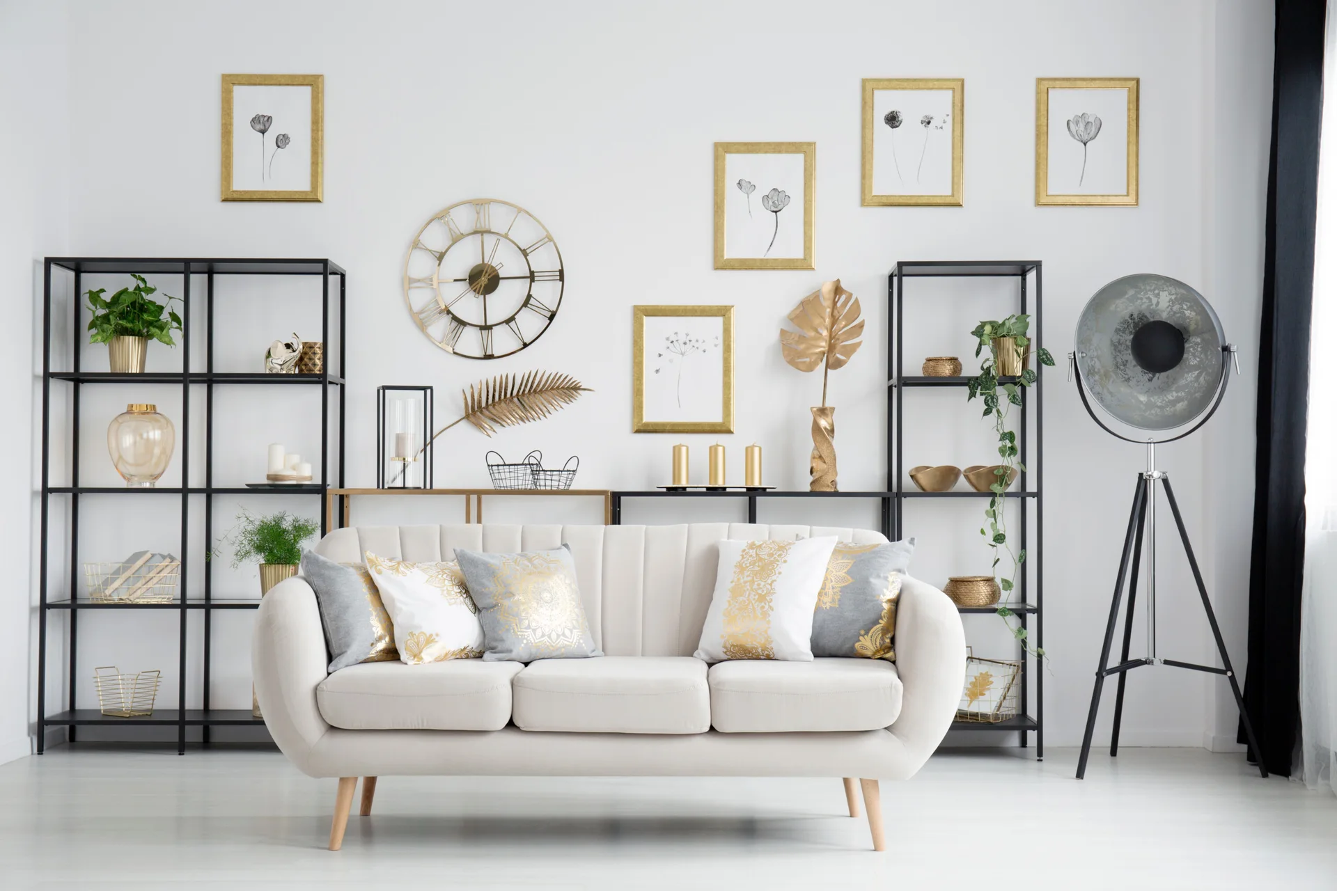 A cream channel-tufted sofa is flanked by two black metal shelving units against a white wall decorated with gold framed art.