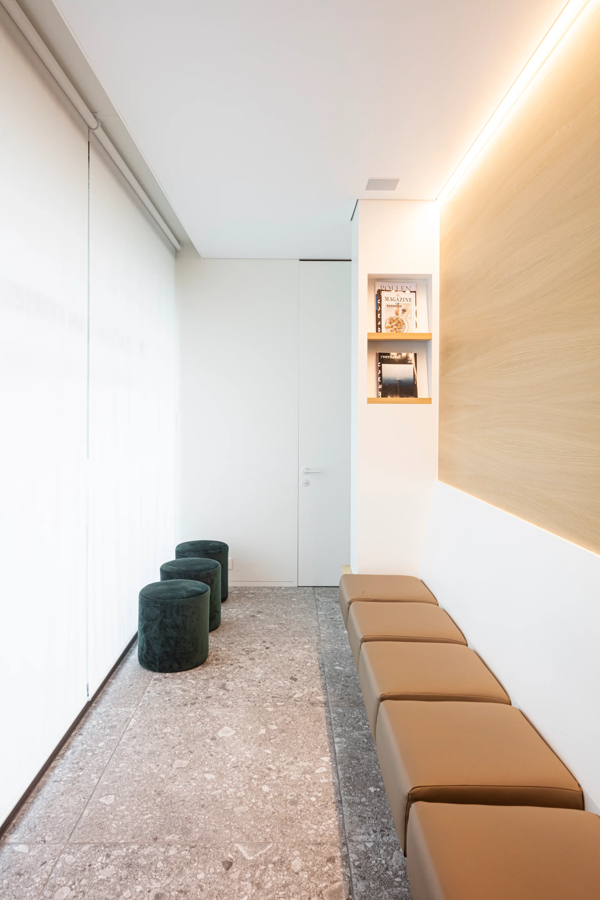 A minimalist hallway featuring a built-in tan leather bench, a row of round green stools, a terrazzo tiled floor, and soft cove lighting.