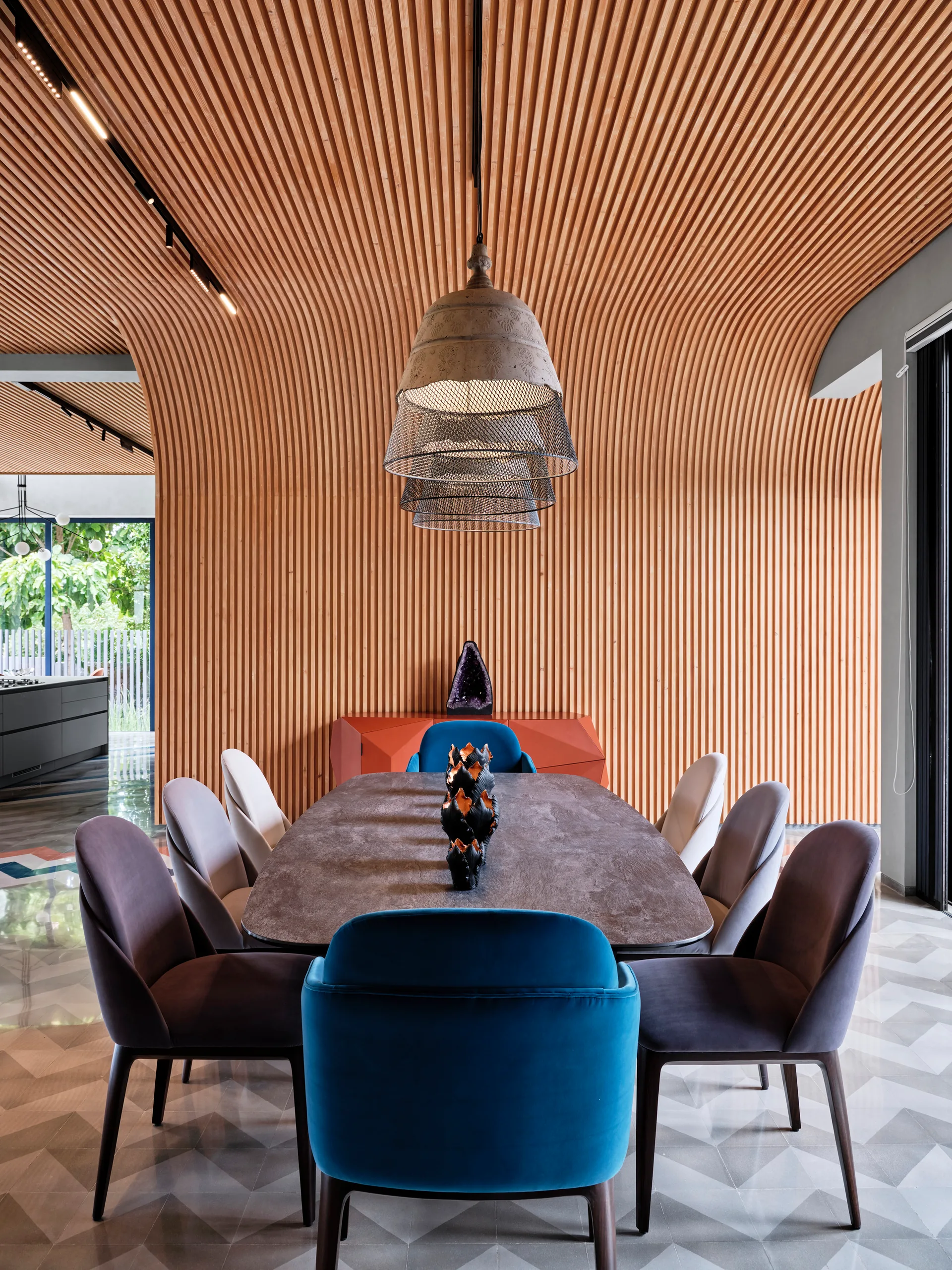 A modern dining table surrounded by blue and purple chairs sits before a curved wood-slatted wall and ceiling in a geometric-tiled room.