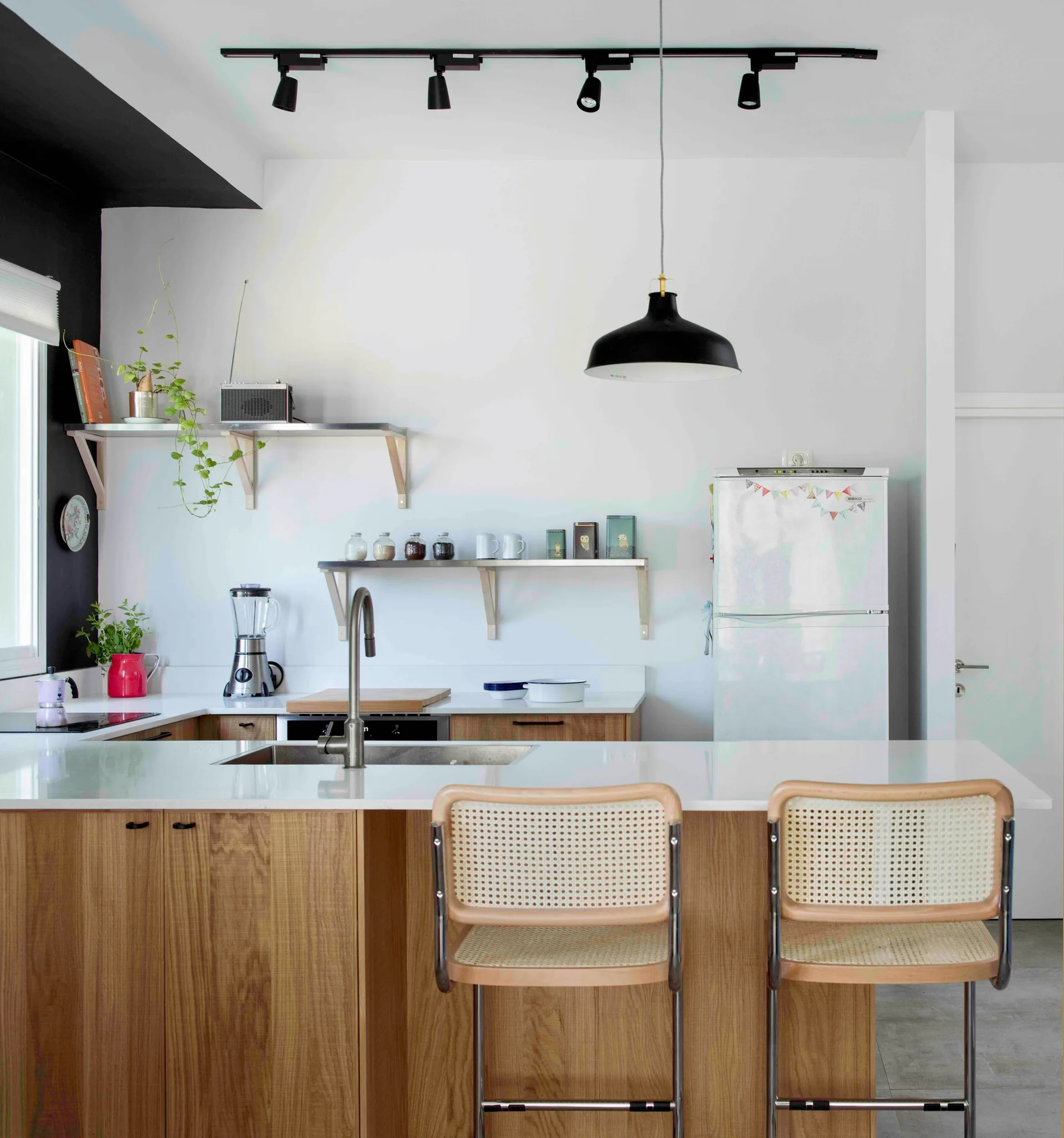 A modern kitchen features a wood-paneled island with a white countertop, two rattan stools, open metal wall shelves, and black track lighting.