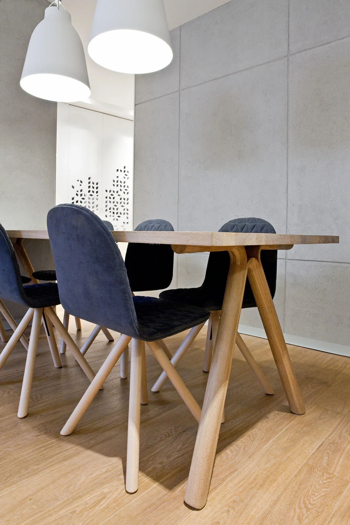 A light wood dining table with dark grey quilted chairs sits in front of a grey paneled wall under white pendant lights.