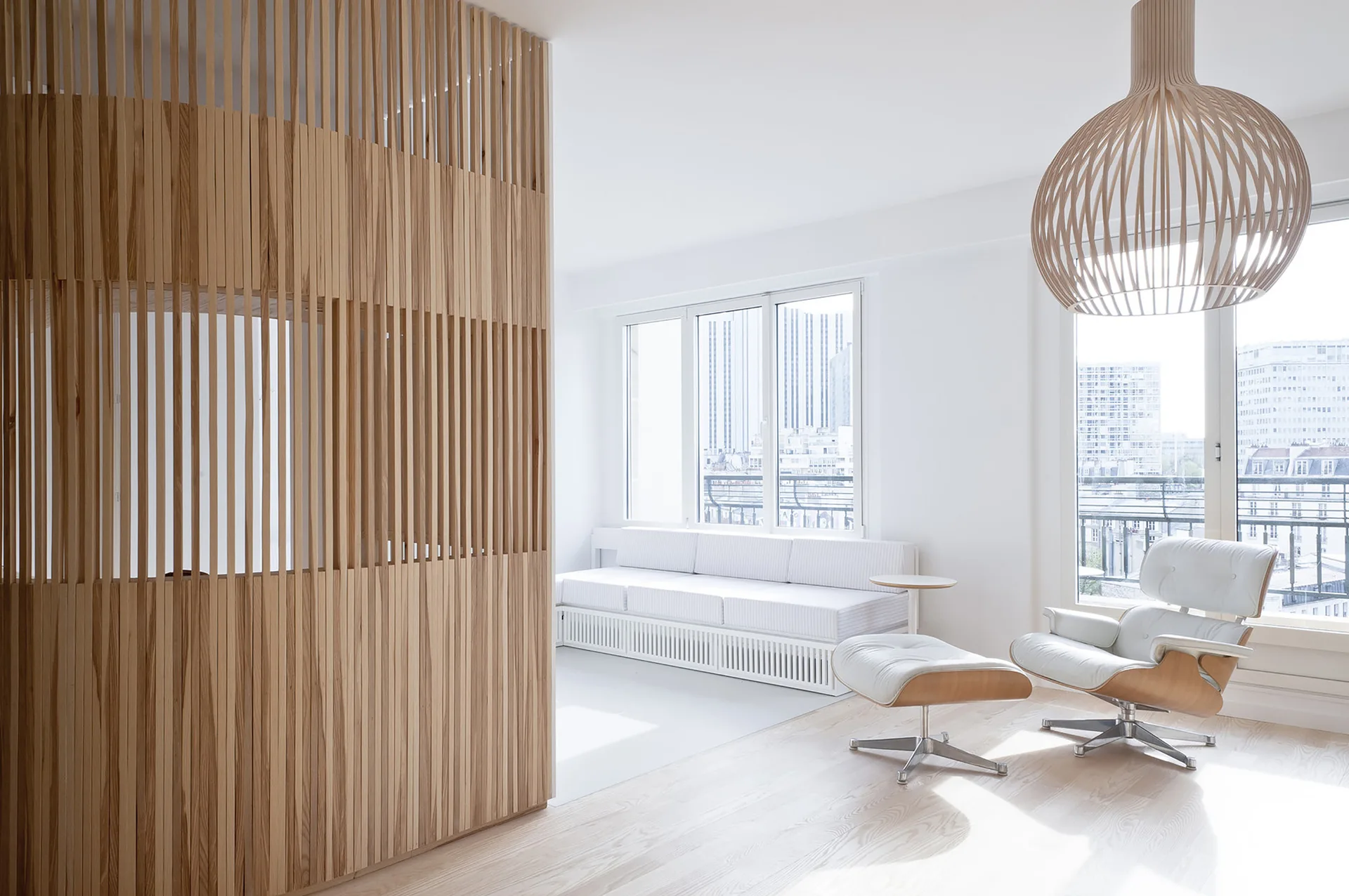 A minimalist white living room features a floor-to-ceiling wooden slat room divider, a white leather lounge chair, and a spherical pendant lamp.