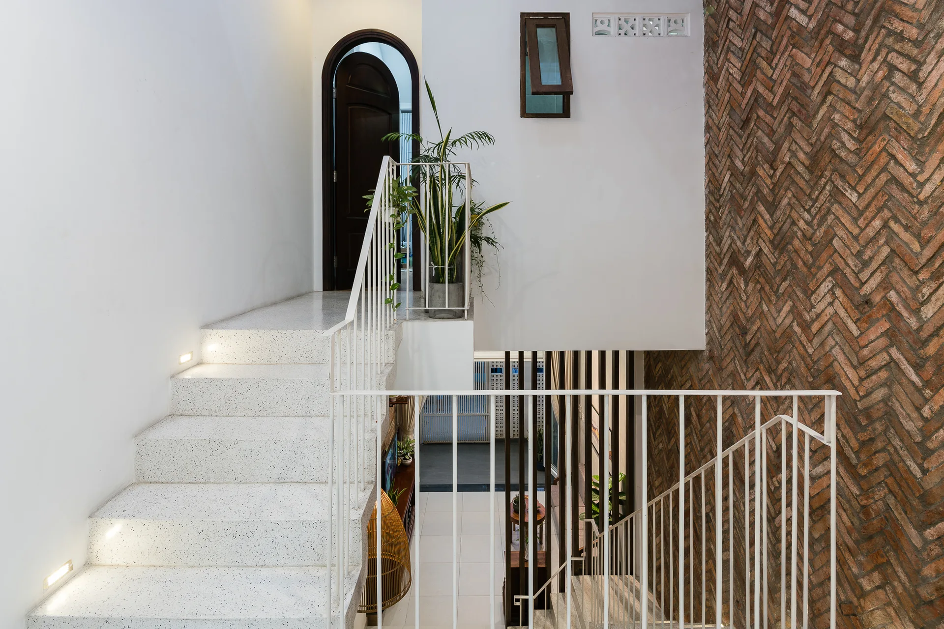 A modern terrazzo staircase with integrated lighting features a white metal railing alongside a tall herringbone-patterned brick accent wall.