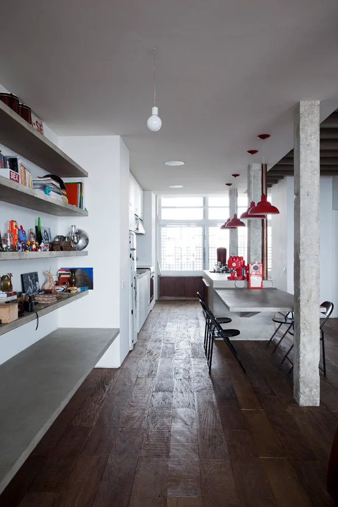 An industrial kitchen features a concrete island, floating concrete shelves, dark wood floors, and bright red pendant lights.