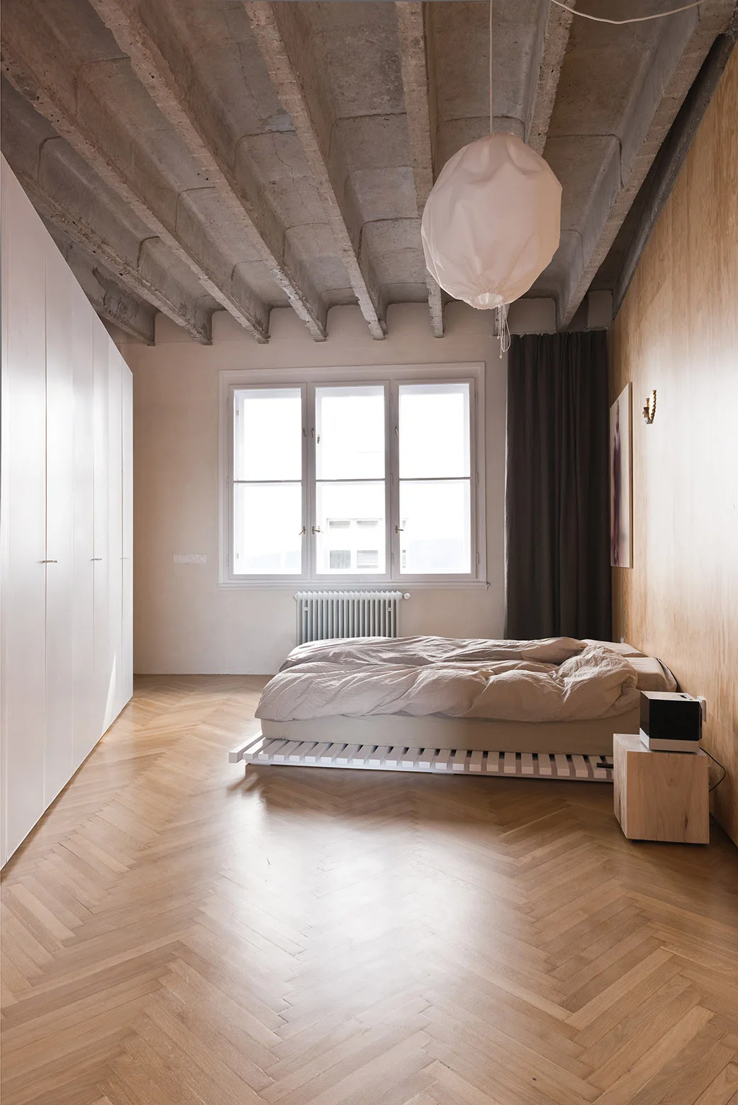 A minimalist bedroom featuring a low platform bed on a herringbone wood floor beneath an exposed concrete beam ceiling.