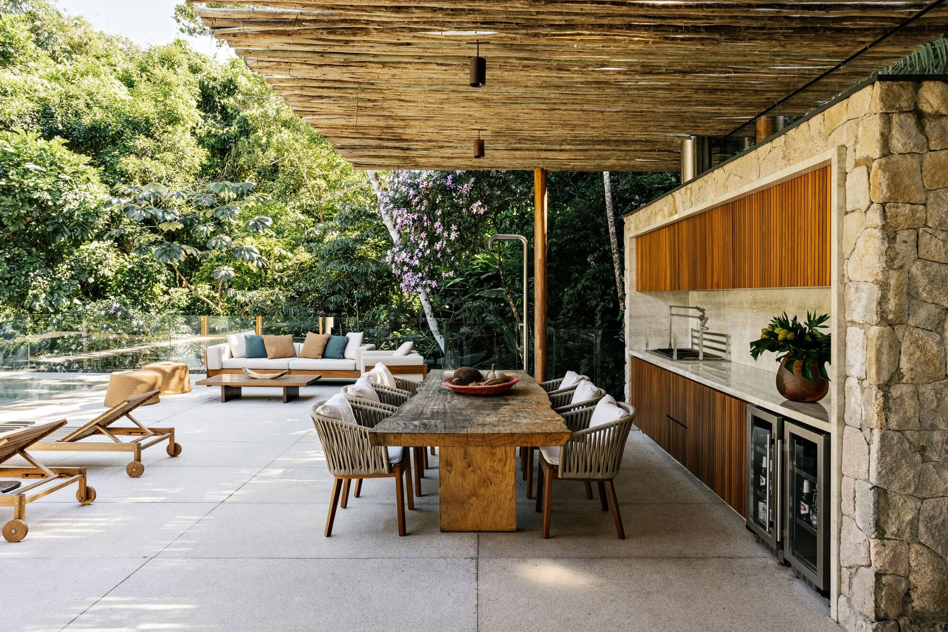 A covered outdoor patio features a rustic wood dining table with woven chairs next to a built-in kitchen against a stone wall.