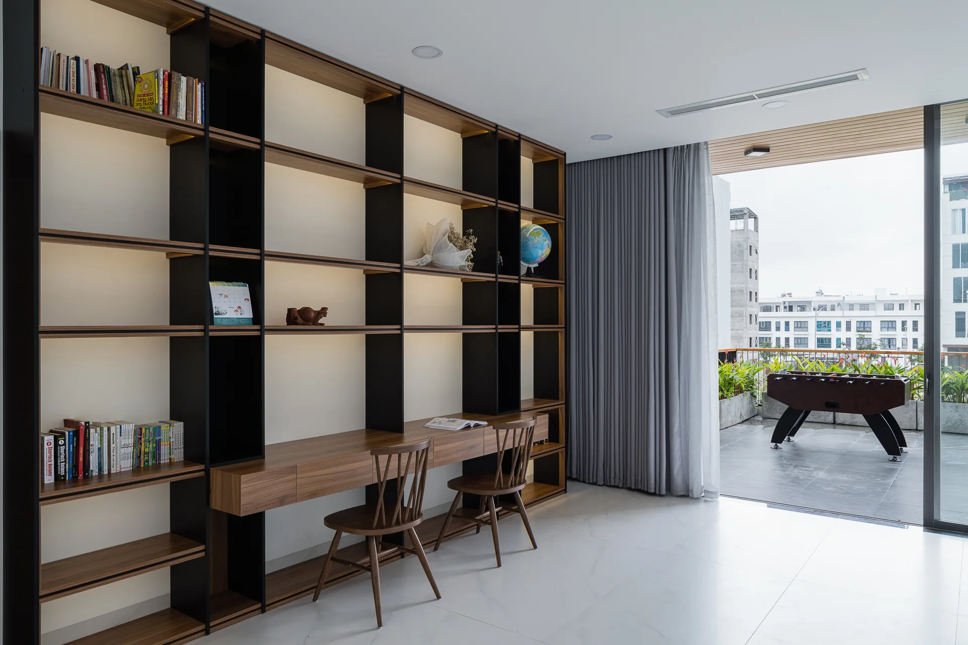 A large wooden built-in shelving unit with an integrated double desk and two spindle-back chairs faces a sliding glass balcony door with grey curtains.