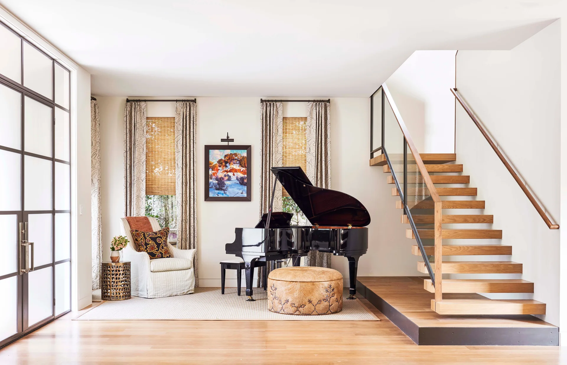 A spacious living room featuring a glossy black grand piano, an open floating wooden staircase, and an upholstered armchair near tall windows.